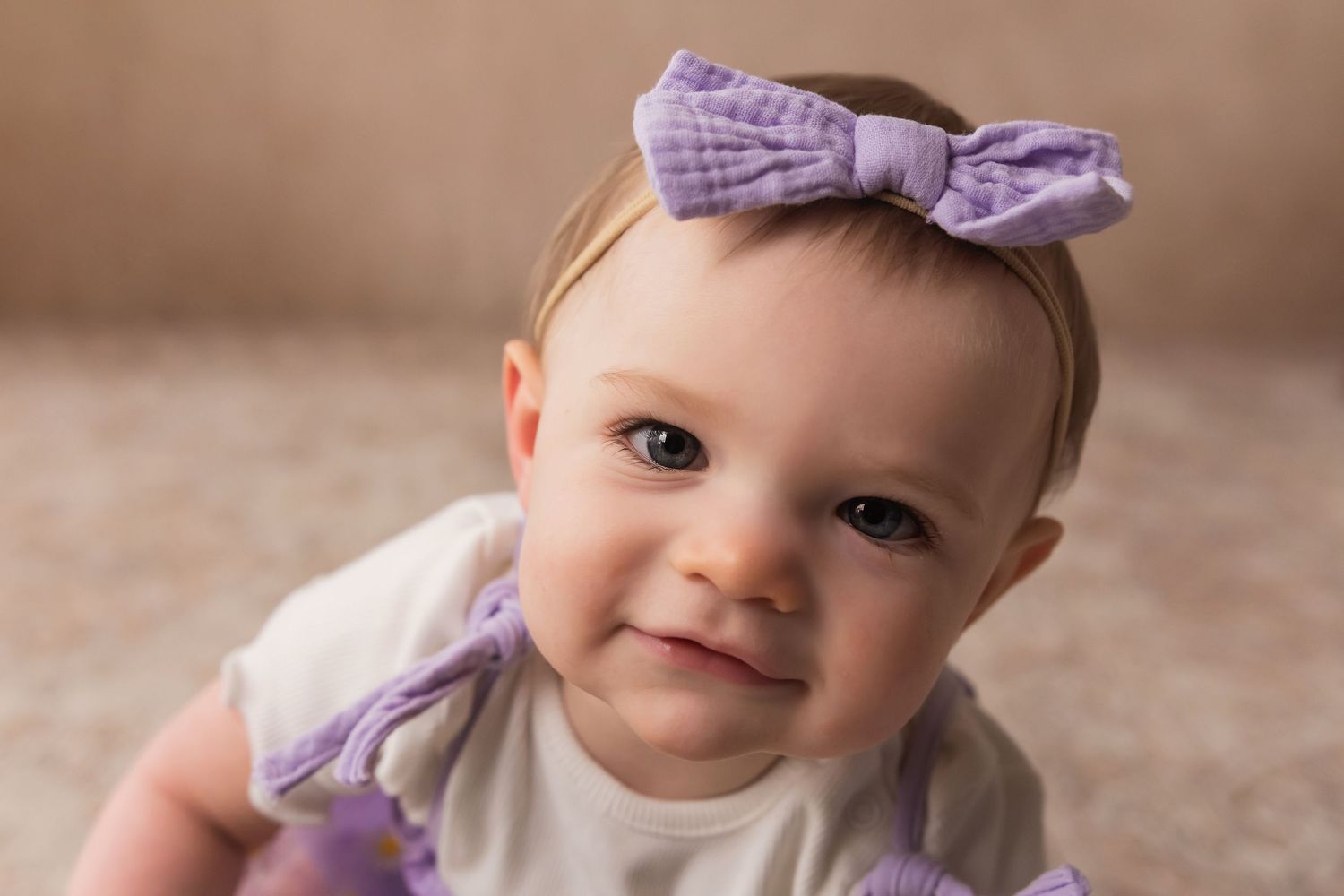 Close-up portrait of a smiling baby girl wearing a purple bow headband and white outfit.