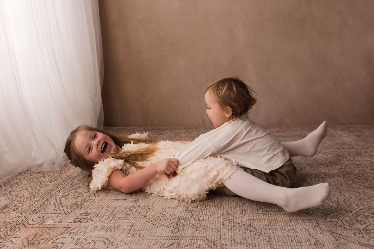 Two young siblings playfully wrestling on a patterned rug in a studio, wearing cream and neutral toned outfits.