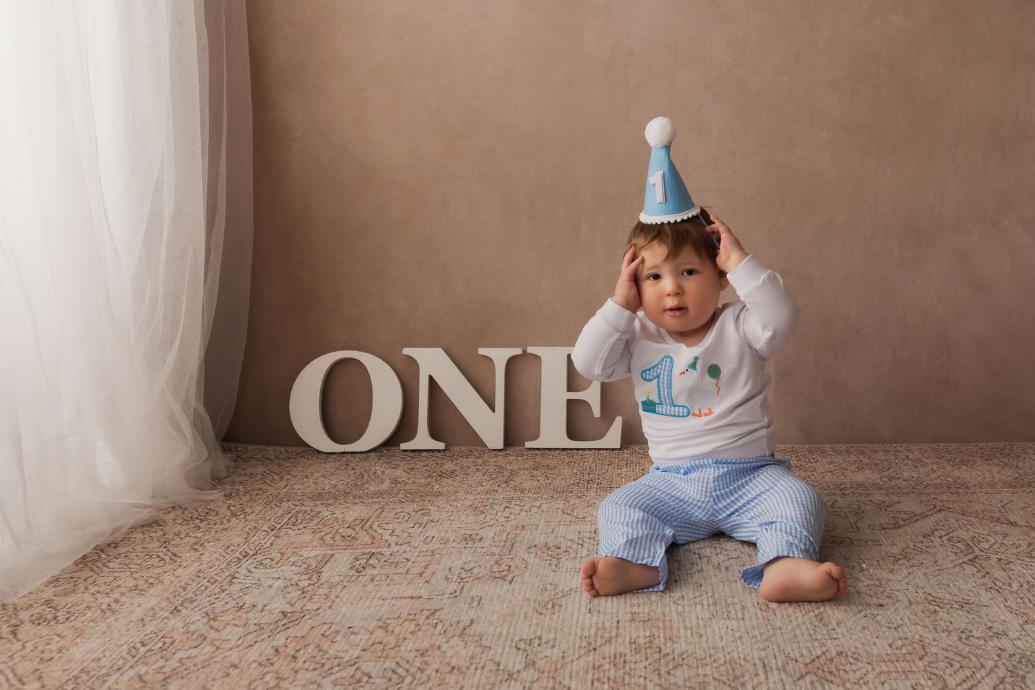 Curious baby boy in blue outfit and party hat sits beside large ONE letters for first birthday.