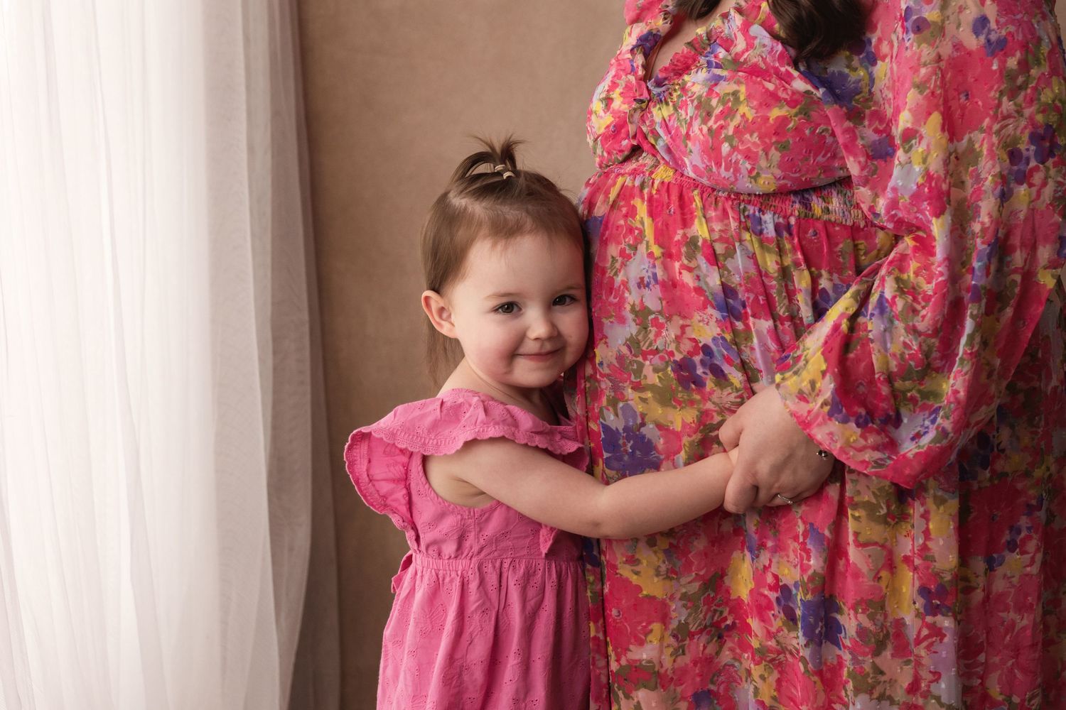 Smiling toddler girl in a pink dress hugging her pregnant mother's baby bump near a window.