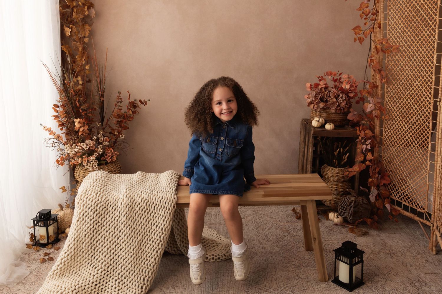 Young girl in denim dress sits smiling on wooden bench with fall autumn decor backdrop.