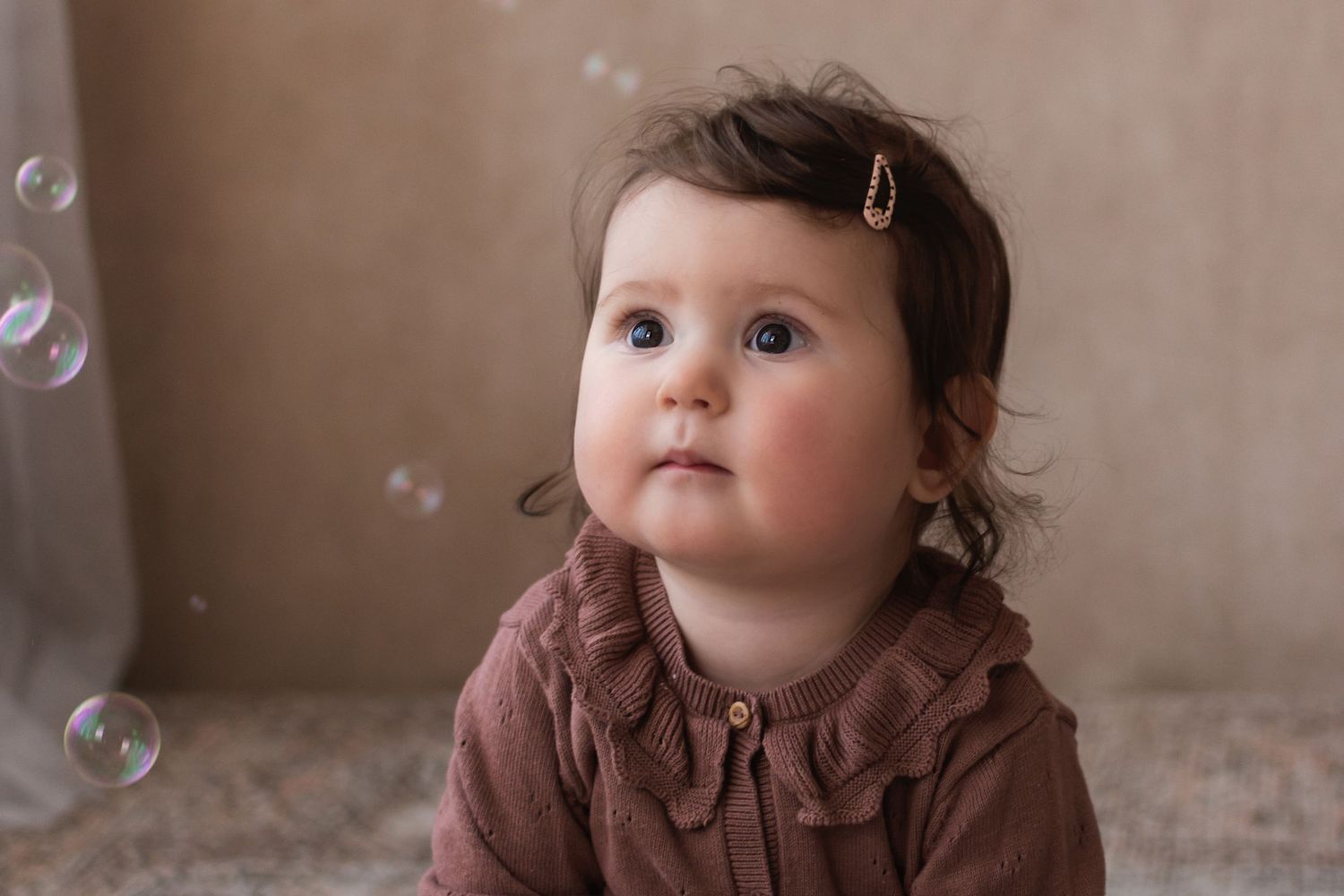 An adorable baby girl with a hair clip looks up curiously at floating soap bubbles.