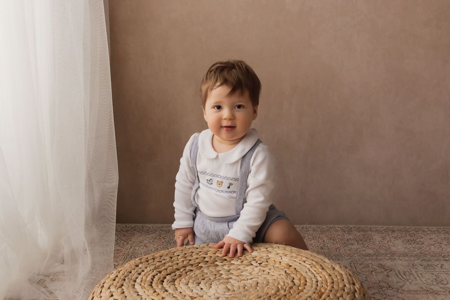 Adorable baby boy in smocked romper sits on a woven round mat beside sheer white curtains.
