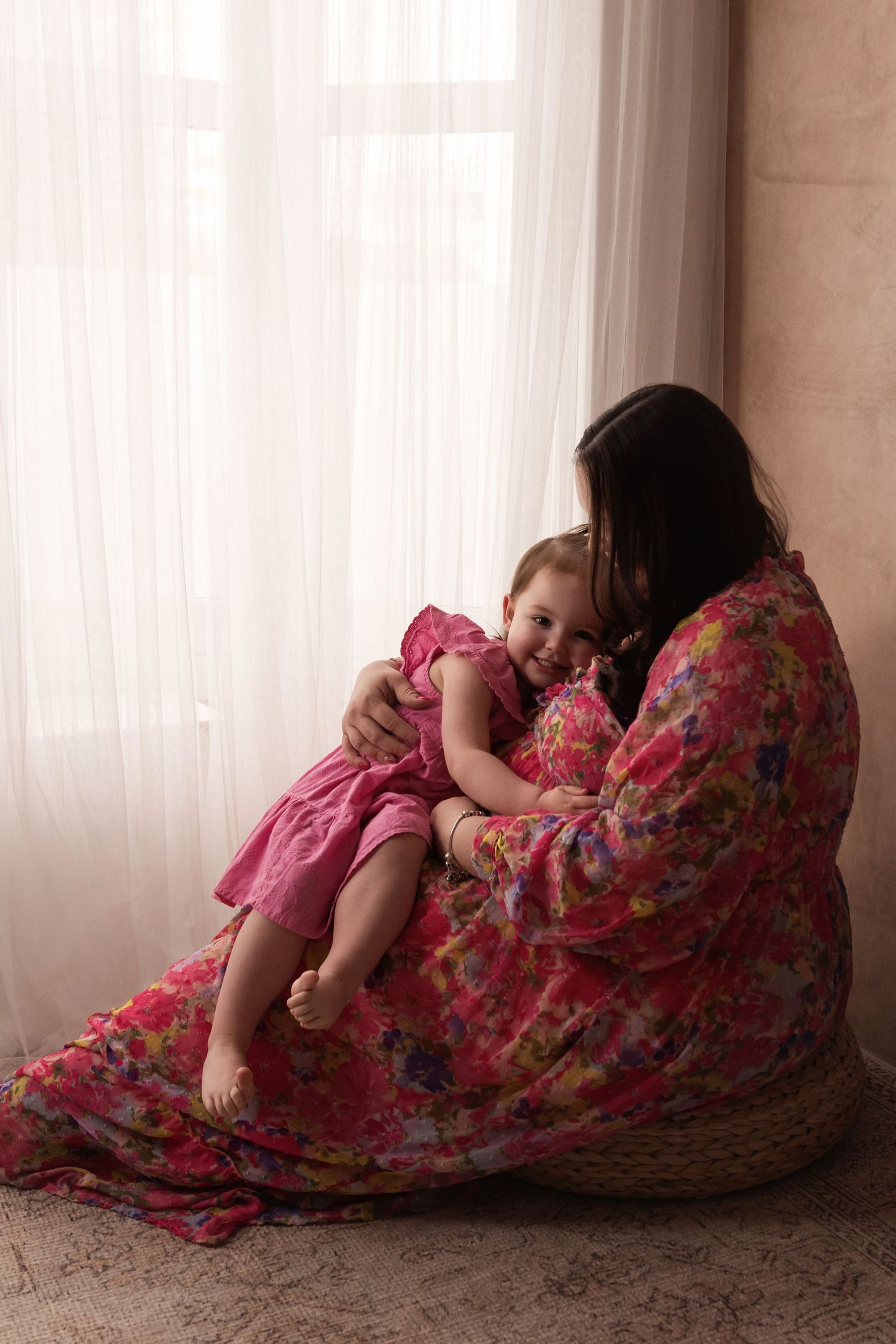 Pregnant woman in a pink floral dress sitting on a pouf cuddling her laughing toddler daughter.
