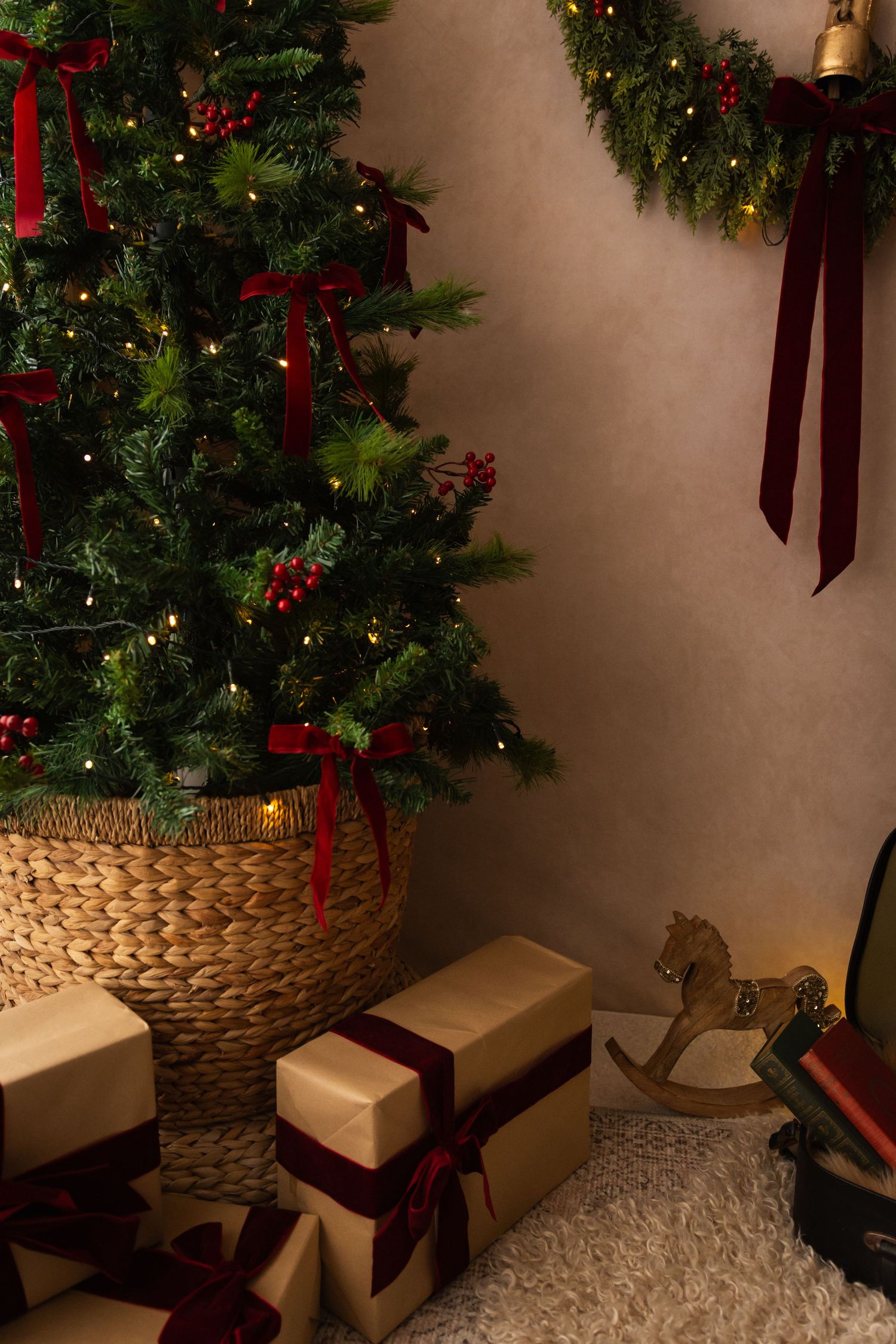 Christmas tree with red bows and lights in wicker basket, surrounded by wrapped gifts and festive garland decor.