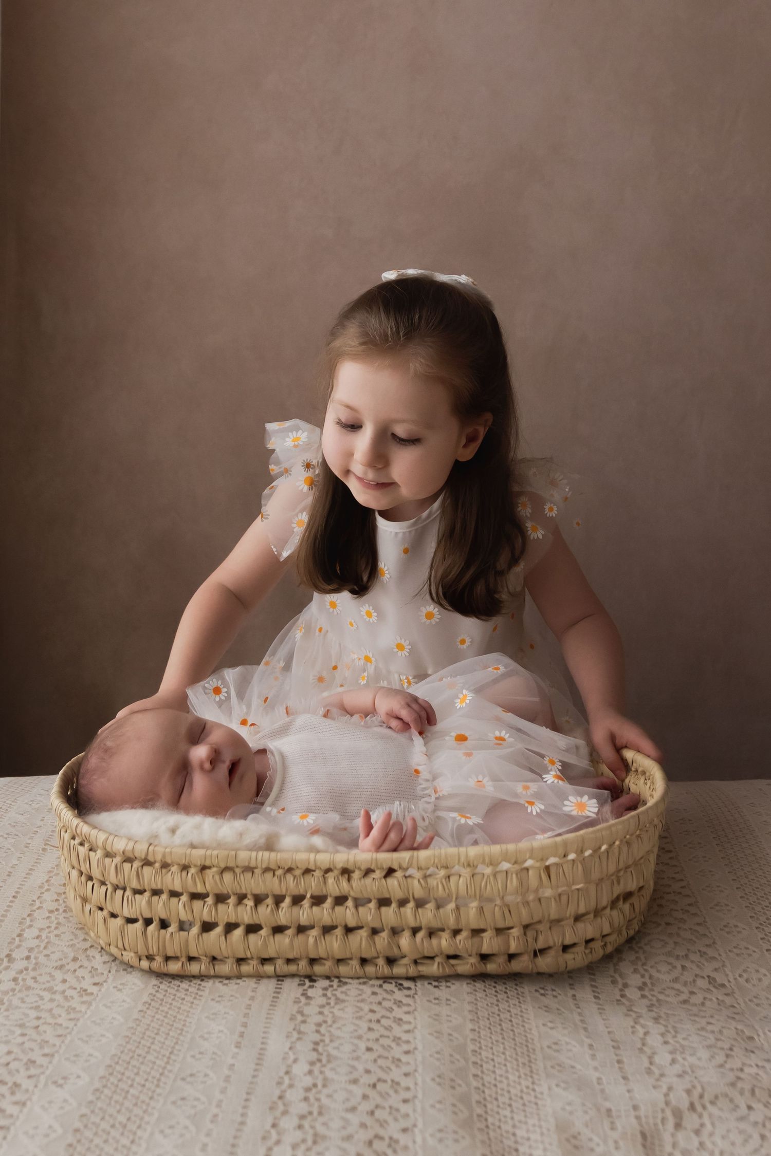 Young girl in daisy dress tenderly gazing at sleeping newborn sibling in wicker basket.