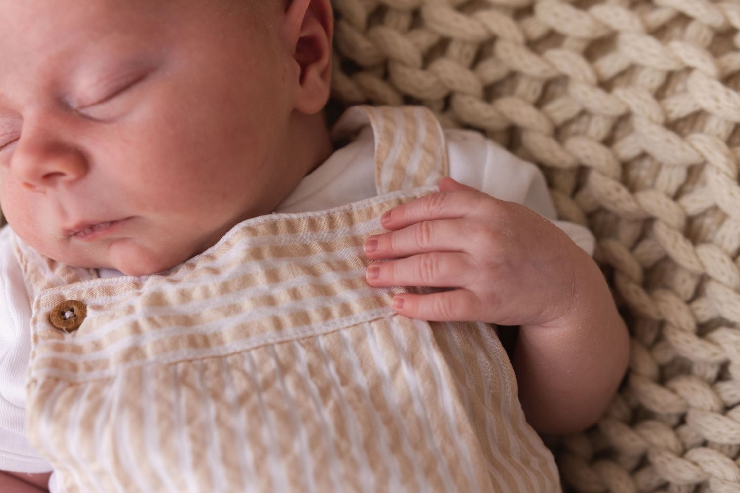 Close-up of sleeping newborn baby in beige patterned romper with tiny hand visible on blanket.