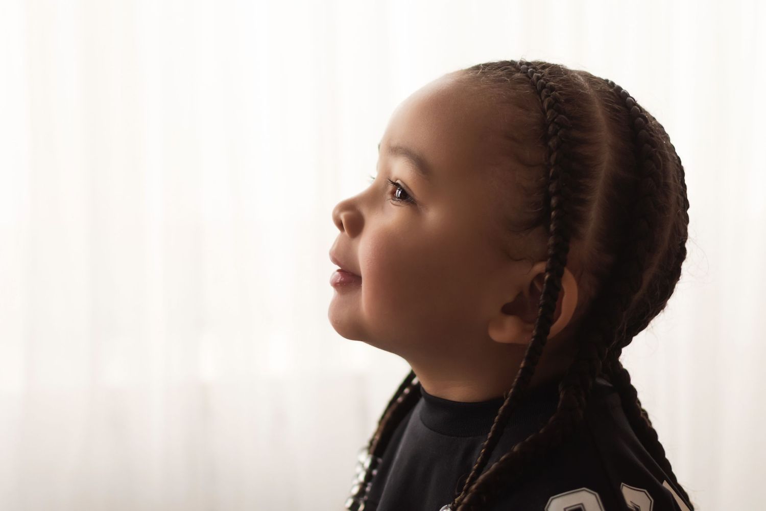 A young child with braided hair gazes upward in a soft profile portrait against a bright white curtain backdrop.