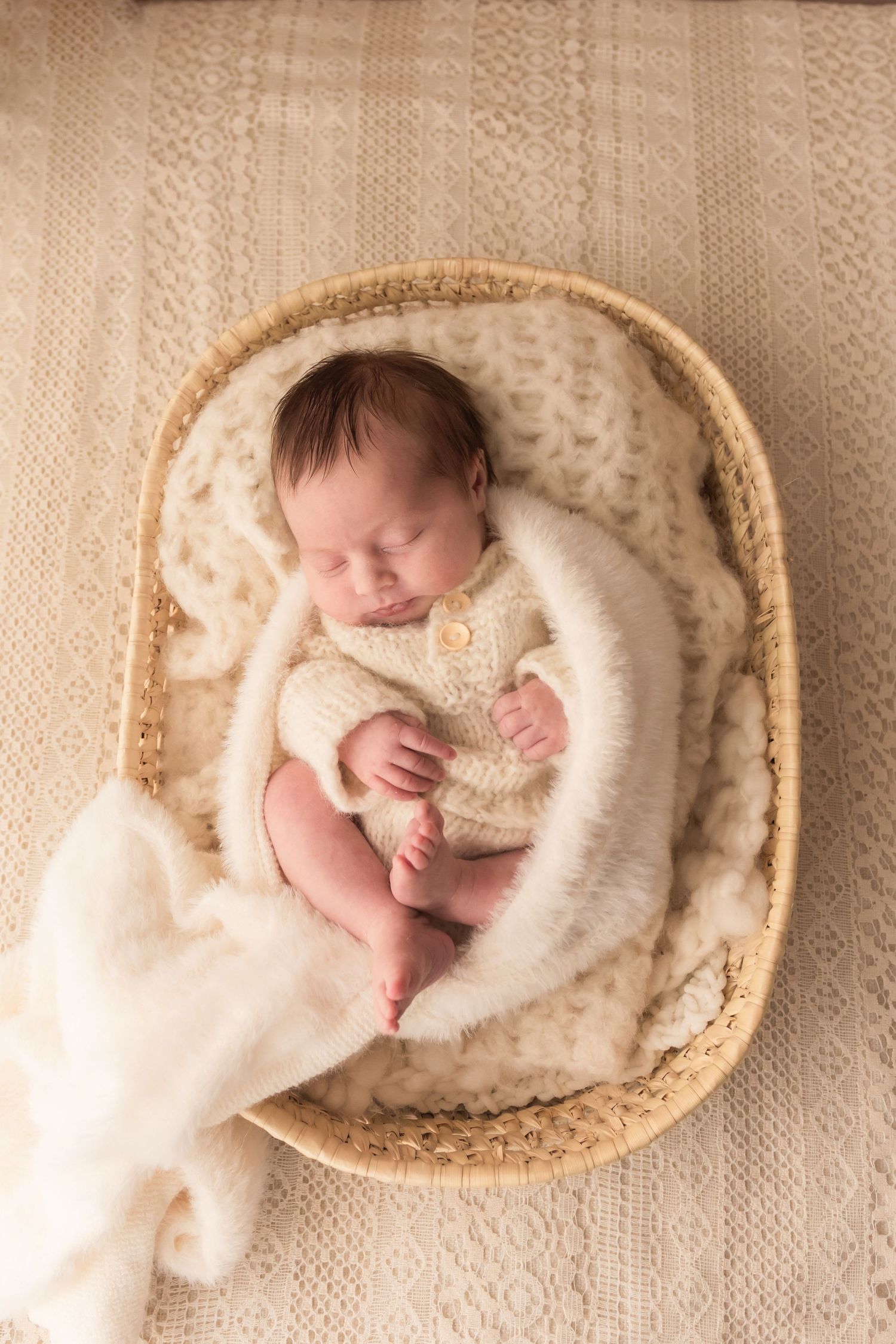Sleeping newborn baby in cream fuzzy romper curled in wicker basket with white fur.