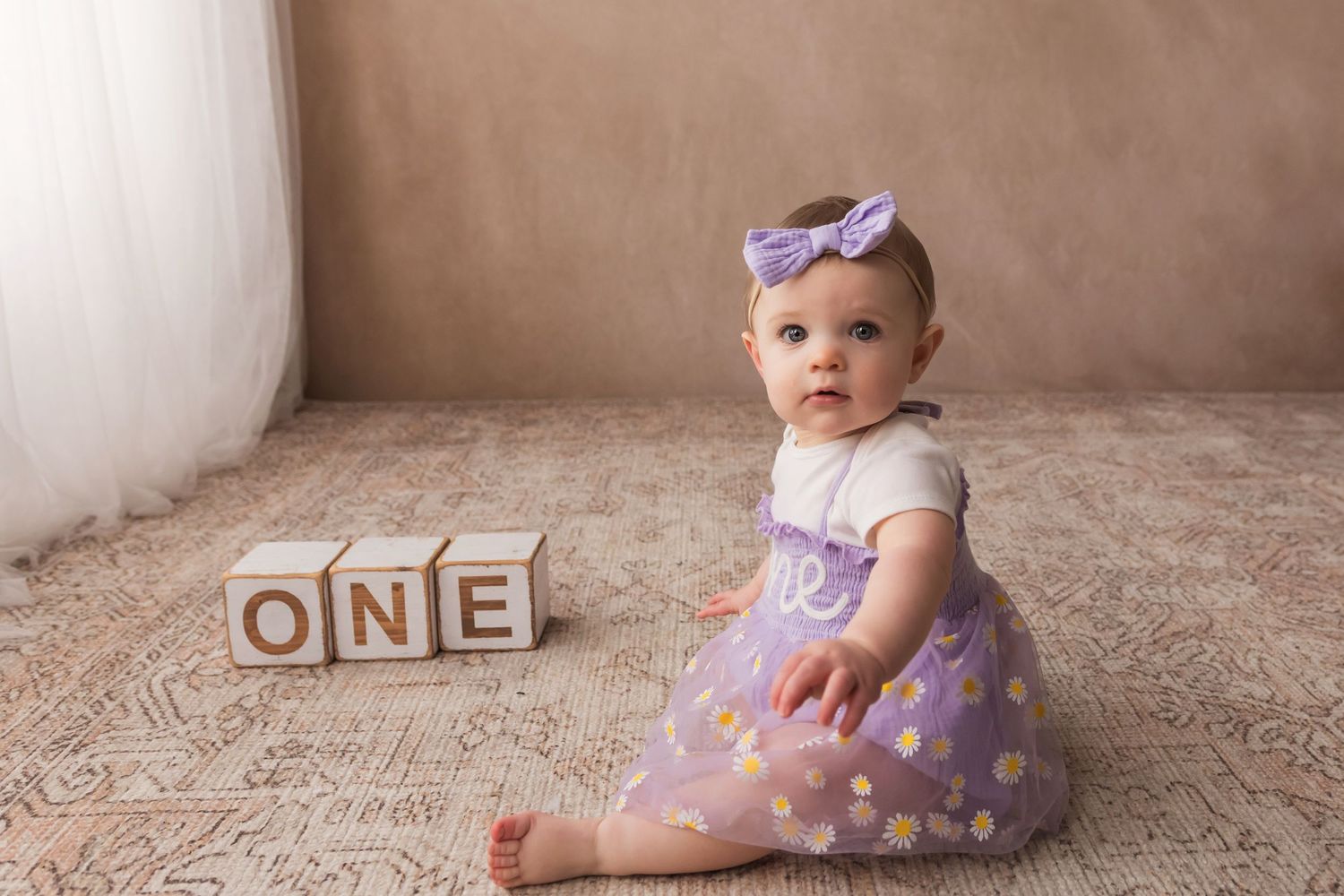 Adorable baby girl in purple polka dot dress sitting beside wooden ONE blocks on first birthday.