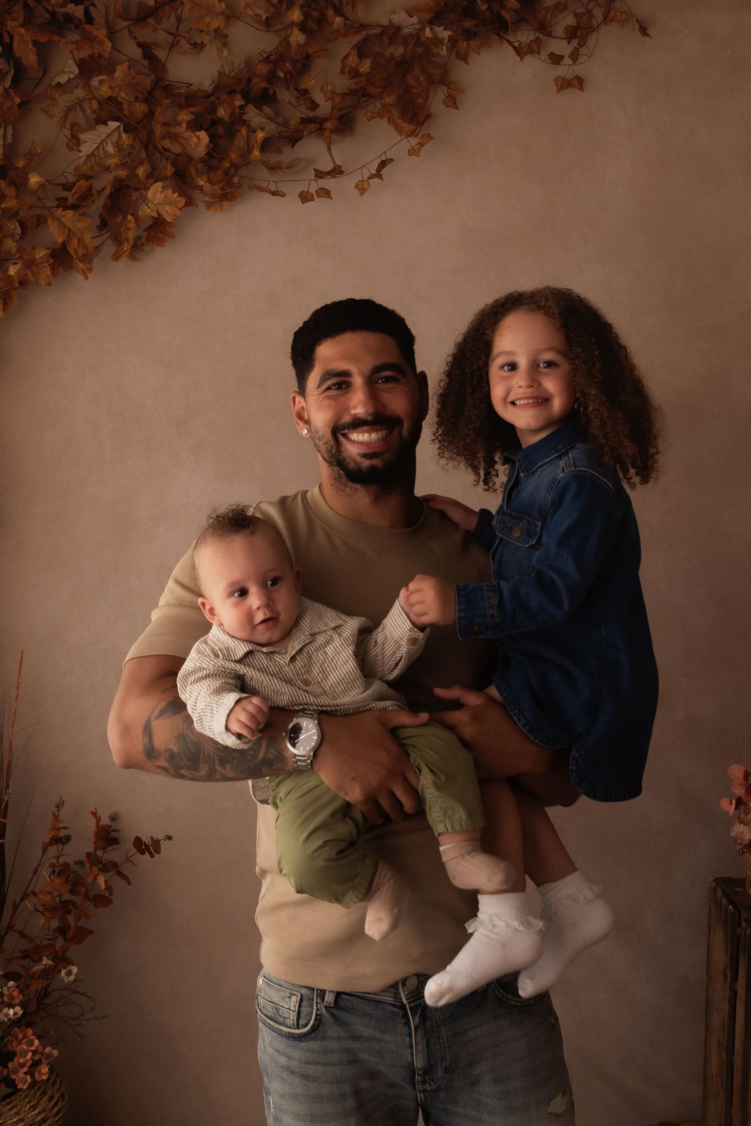 A smiling man holds a baby and a curly-haired toddler girl in a warm autumn-themed studio portrait.