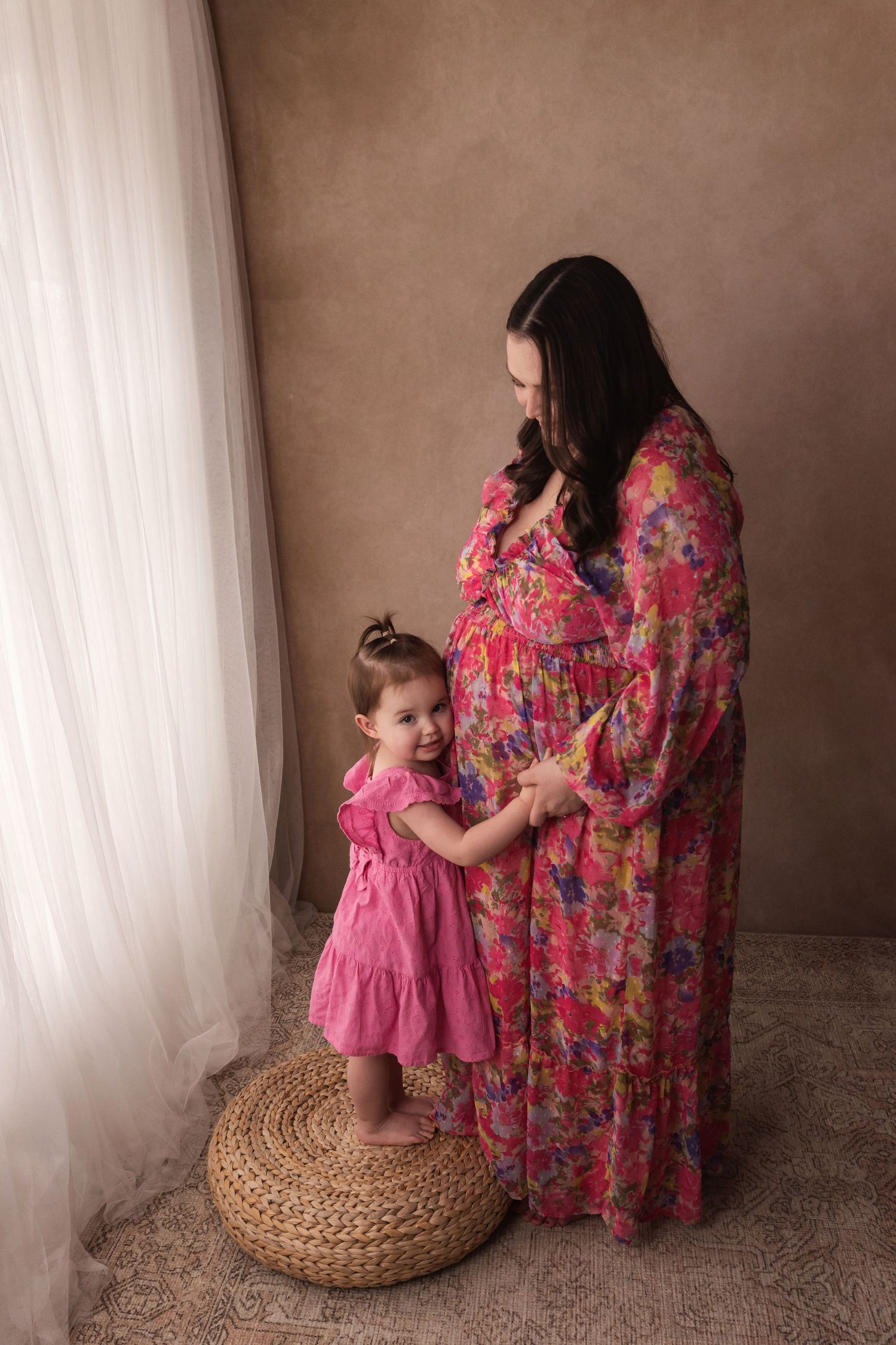 Pregnant woman in a vibrant pink floral maxi dress standing with her toddler daughter by a window.