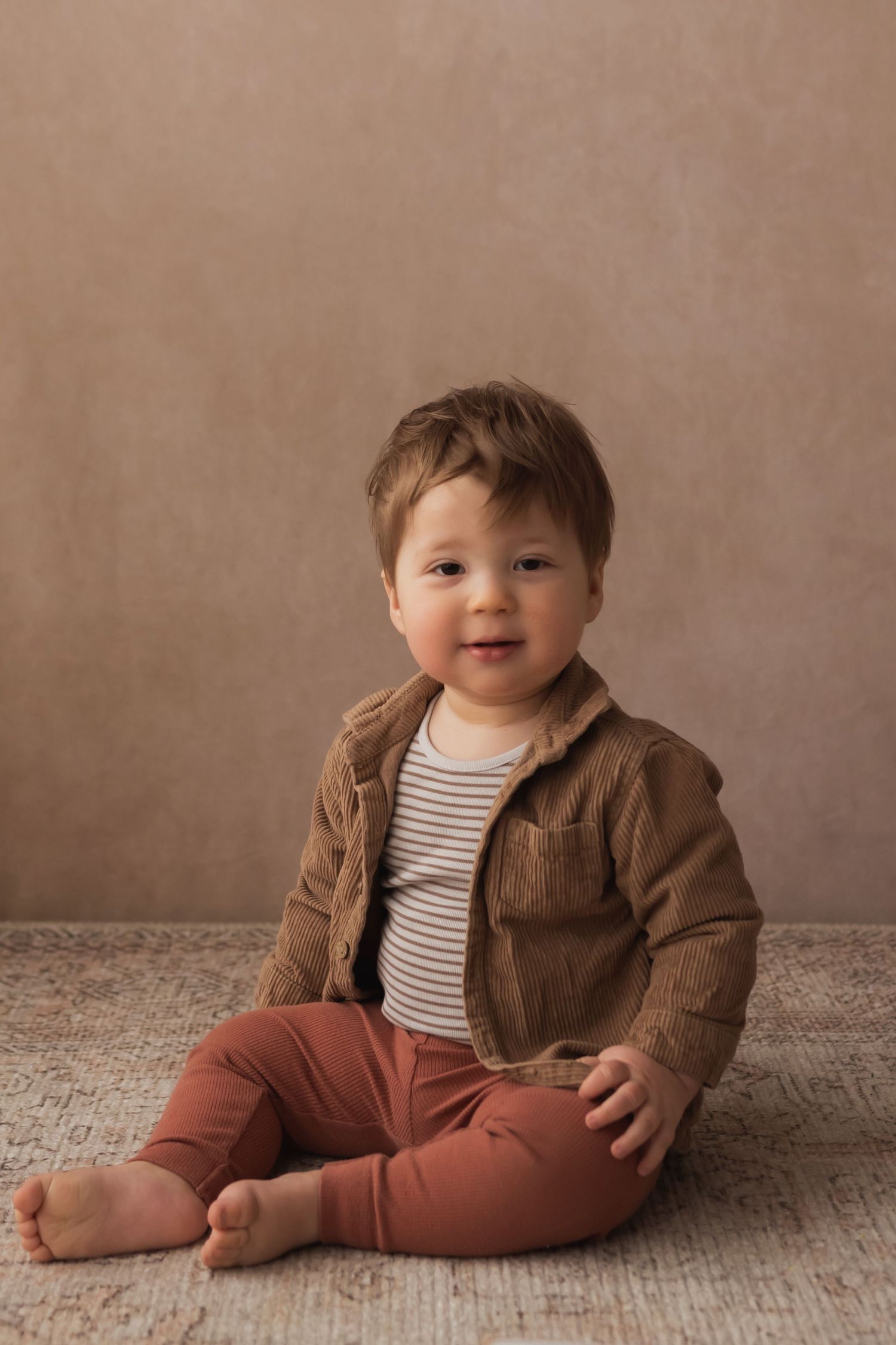 Sweet baby boy in brown jacket and rust pants sits on a rug smiling softly at the camera.