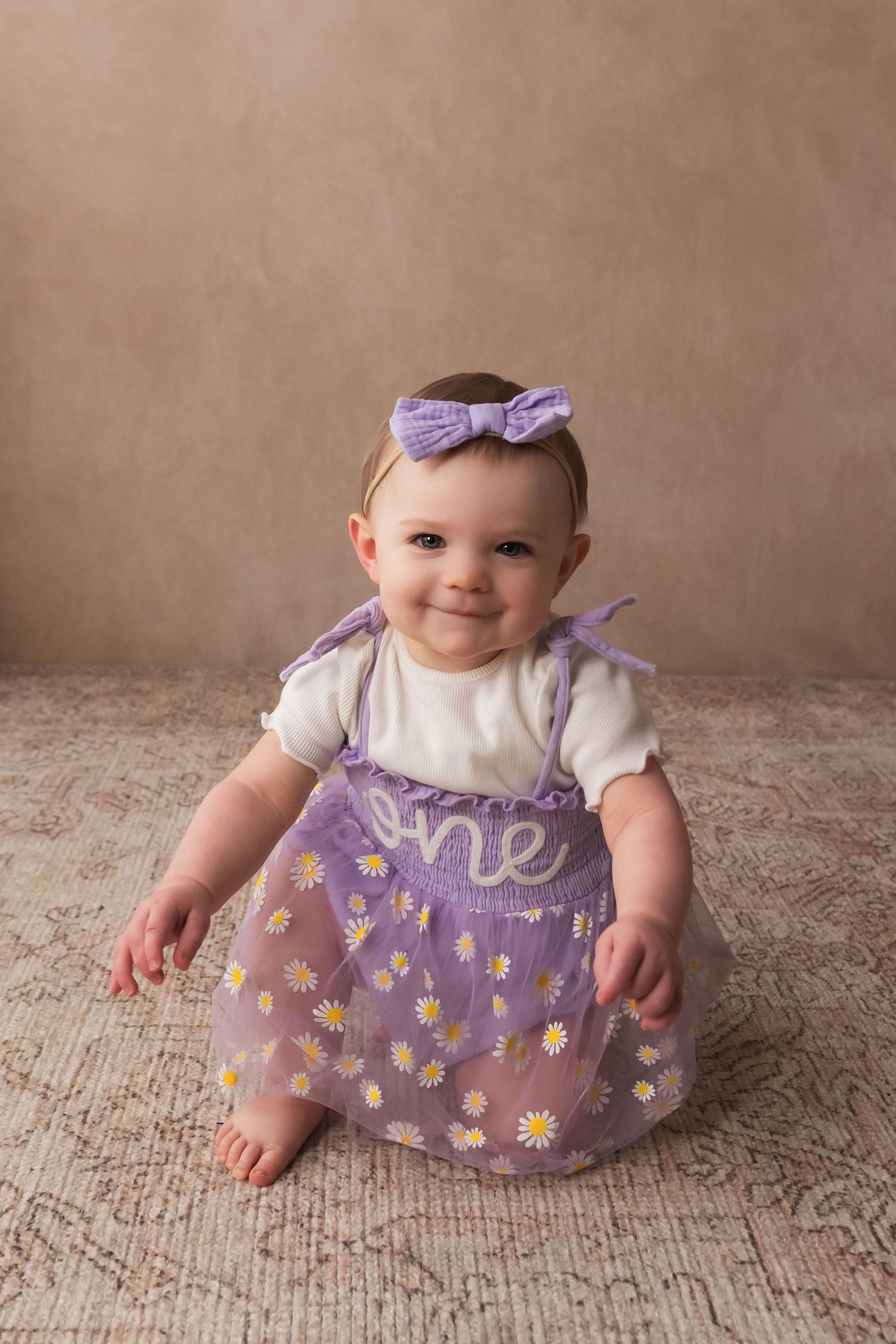Smiling baby girl in purple daisy ONE dress and bow headband celebrates her first birthday.
