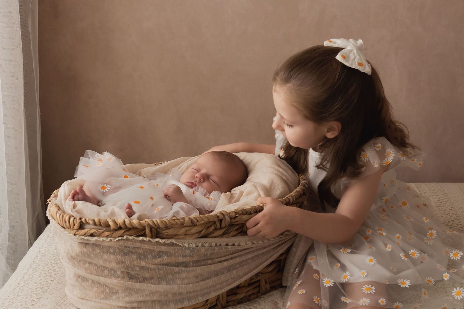 Big sister in floral dress lovingly kissing newborn baby lying in a wicker basket.
