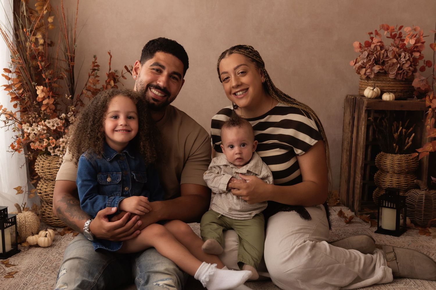 Family of four posing for a warm autumn-themed studio portrait with fall decor, pumpkins, and dried flowers in the background.