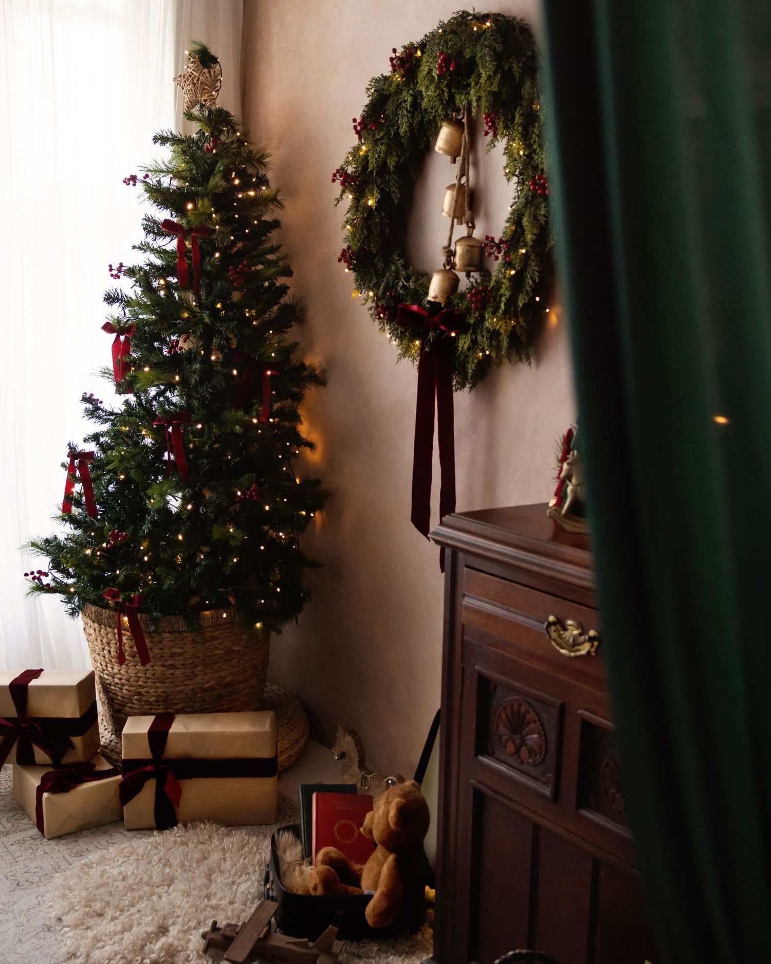 Cozy Christmas corner with decorated tree, wrapped gifts, teddy bear, and festive wreath on wall beside wooden dresser.