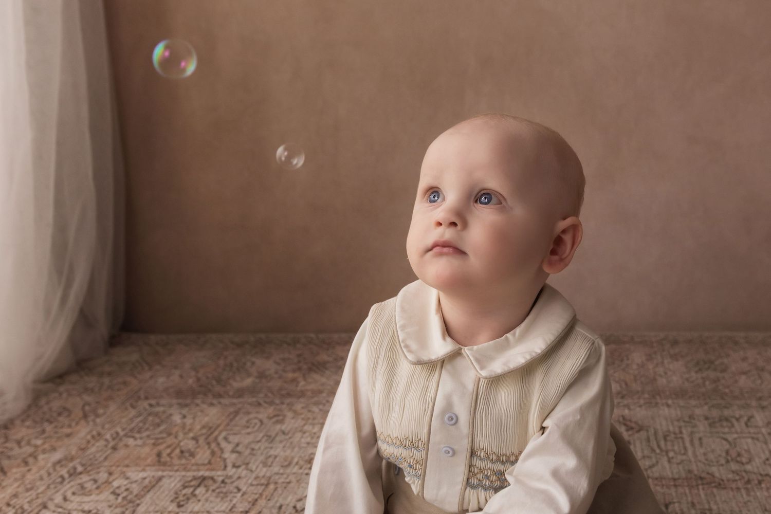 Blue-eyed baby in cream smocked outfit gazes up at floating soap bubbles indoors.