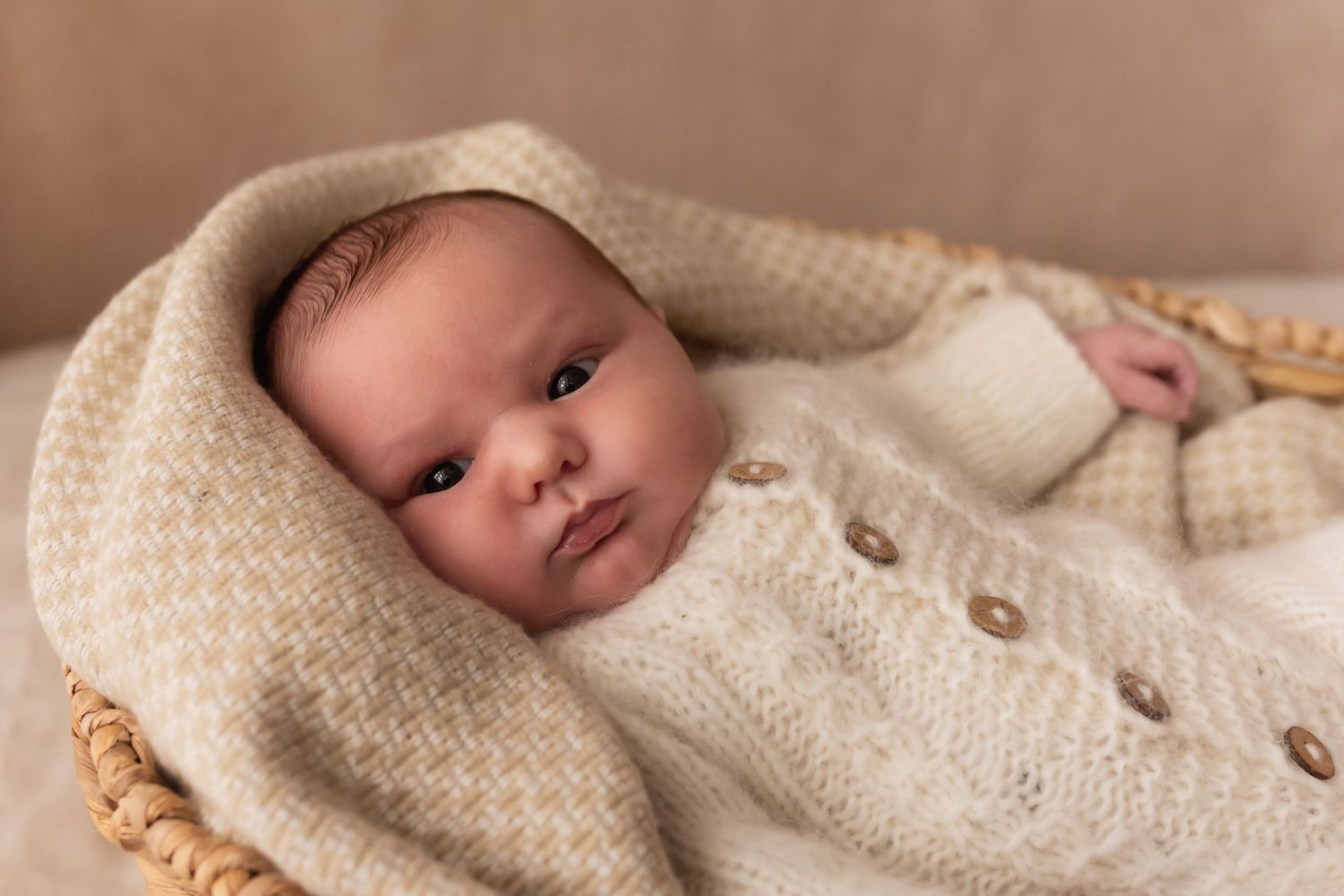 Wide-eyed newborn baby in cream button cardigan wrapped in blanket inside wicker basket.