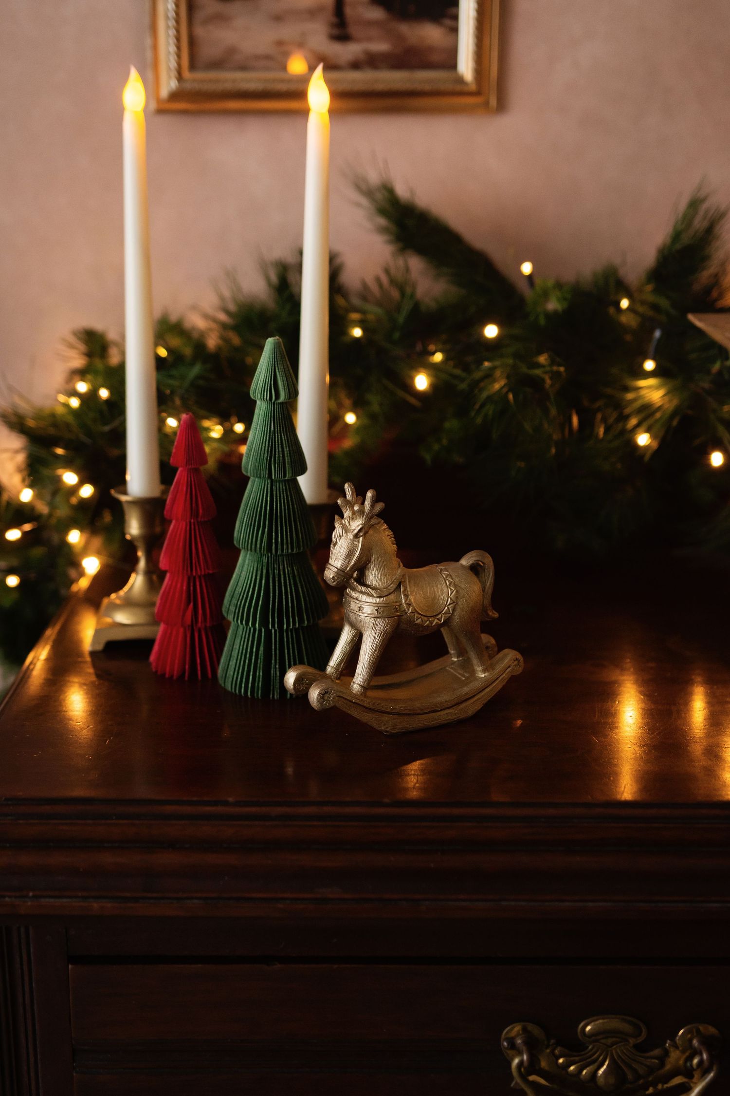 Christmas decor with rocking horse, paper trees, candles, and fairy lights on a dark wooden dresser.