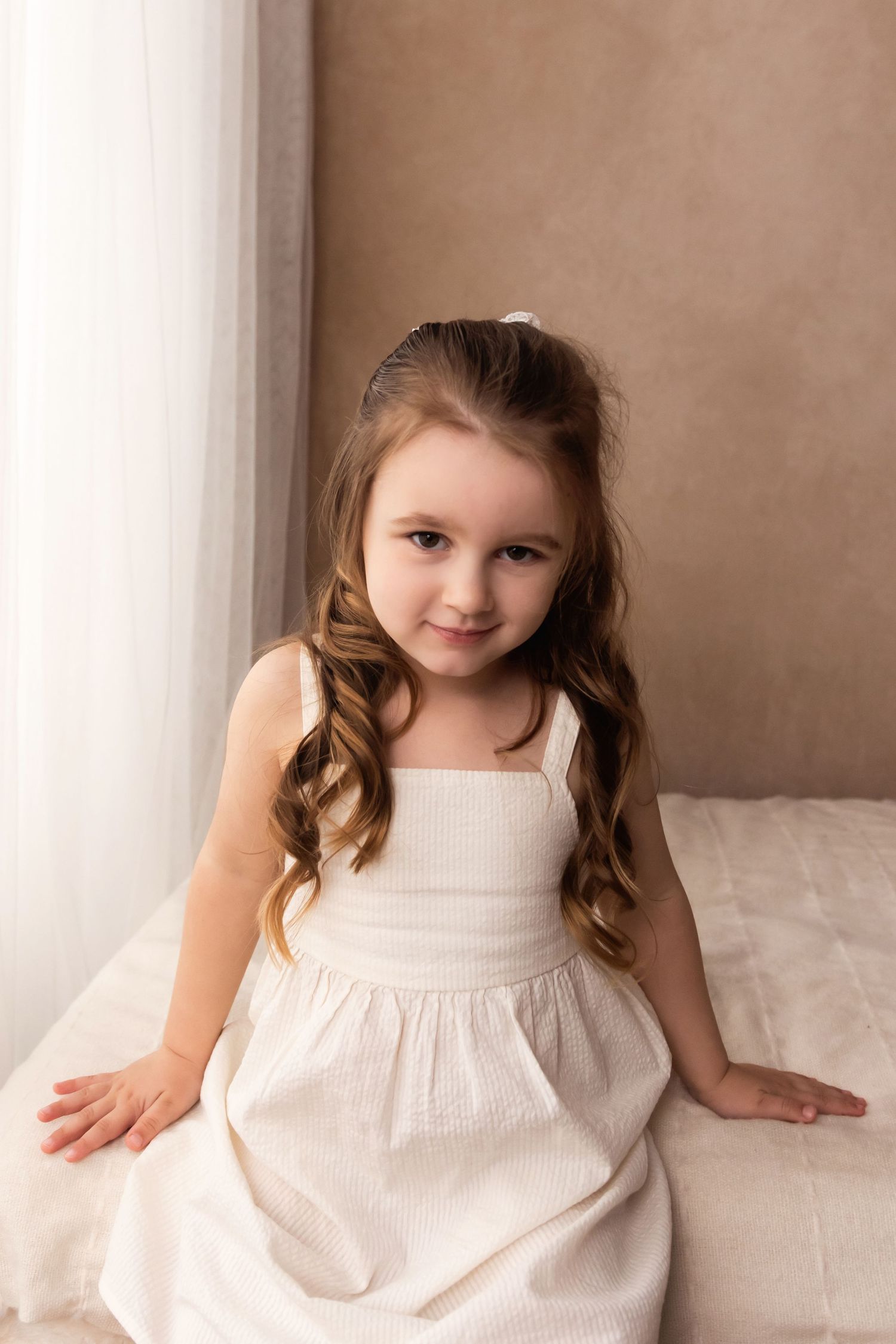 A young girl with long curly hair wearing a white dress sits gracefully, smiling softly in a warmly lit room.