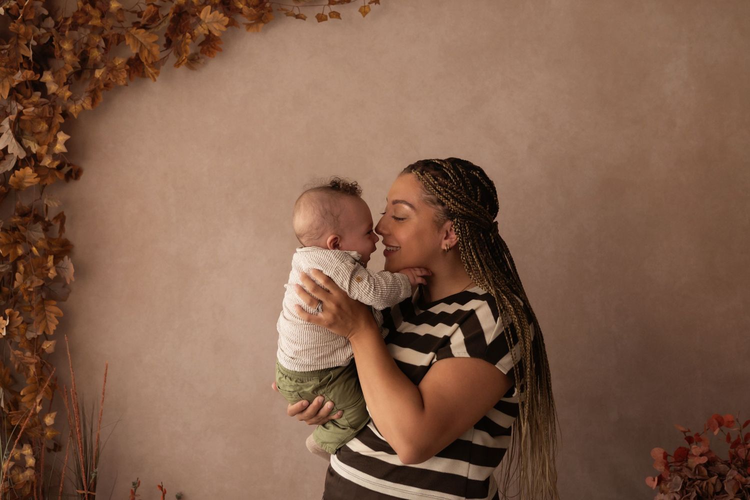 A mother with braided hair kisses her baby against a neutral backdrop with autumn leaves.