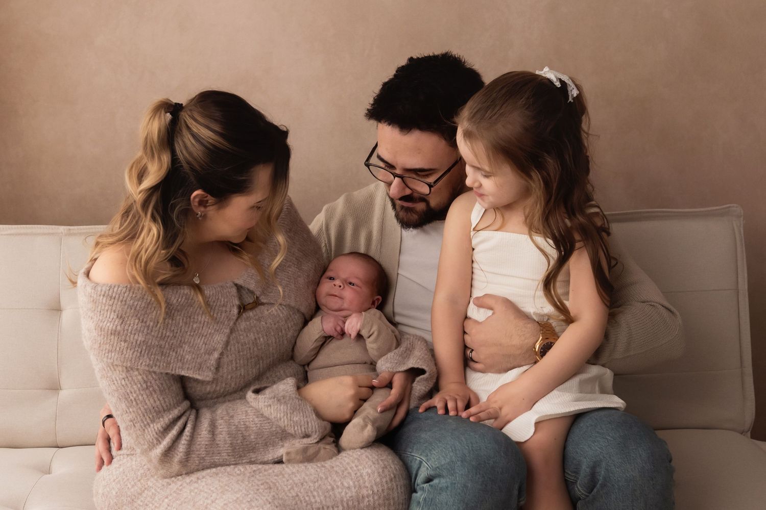 Happy family of four with parents and young daughter admiring newborn baby on sofa.