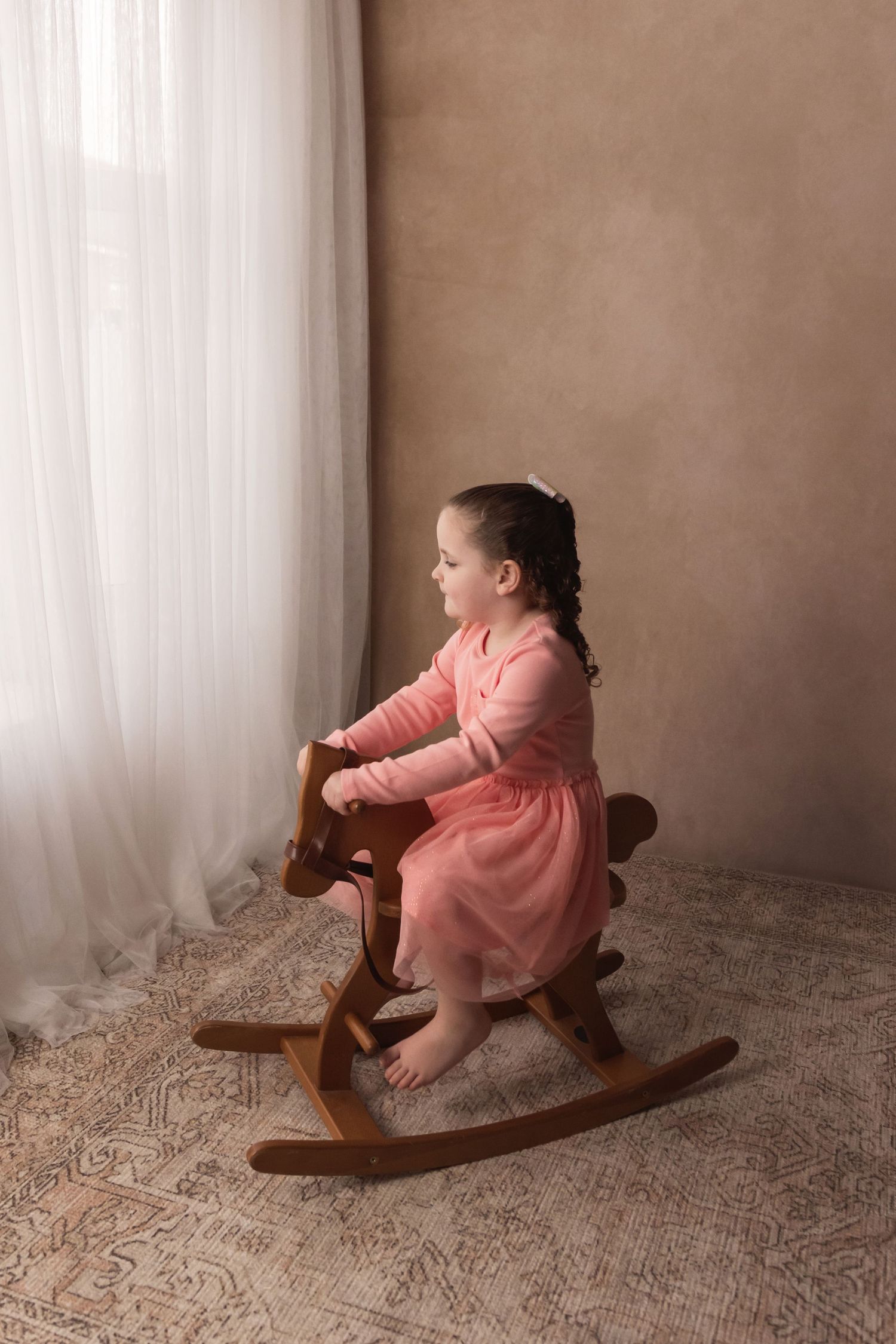 Little girl in pink dress sitting on wooden rocking horse looking out window during photo session.