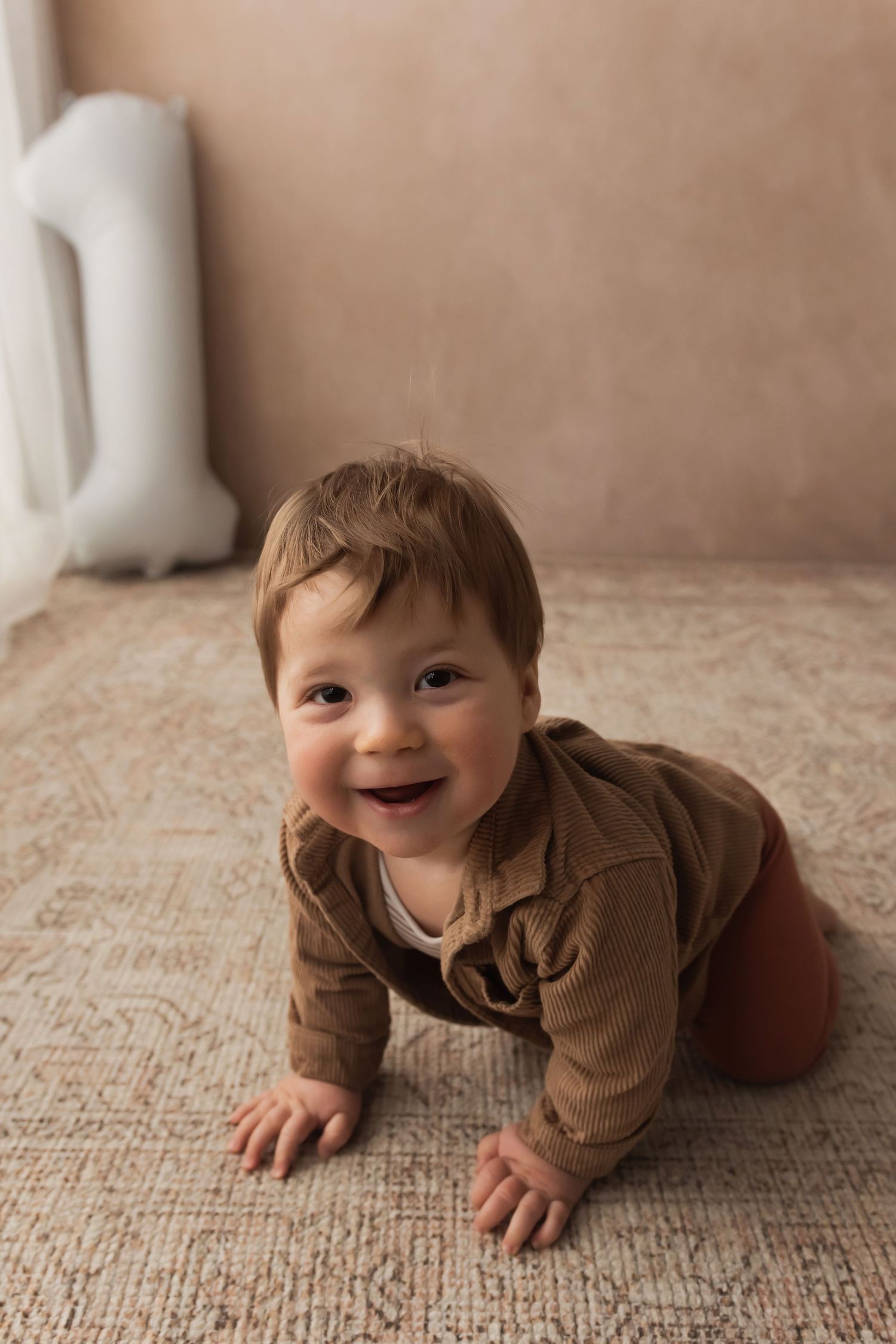 A happy baby boy in a brown outfit crawls and smiles on a patterned carpet.