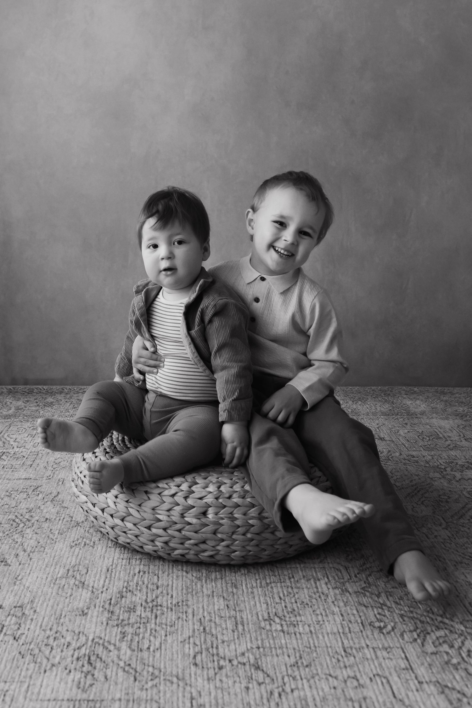 Black and white studio portrait of two young boys sitting together on a woven pouf.