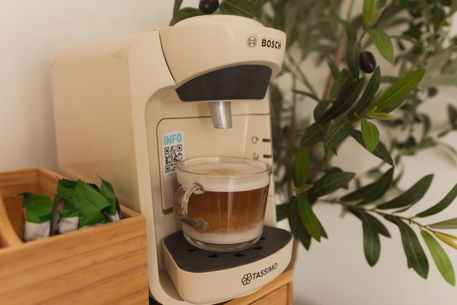Bosch Tassimo coffee machine brewing a latte in a glass cup on a white countertop with green plants.