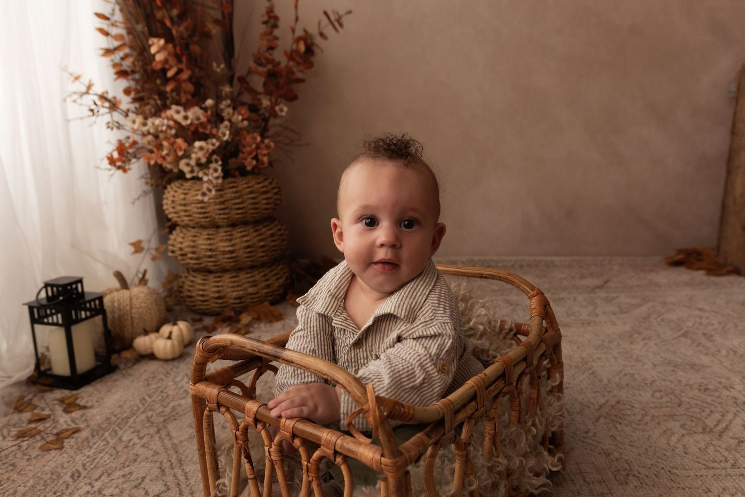 A smiling baby sits in a wicker cradle surrounded by autumn decor, pumpkins, and dried florals in warm tones.