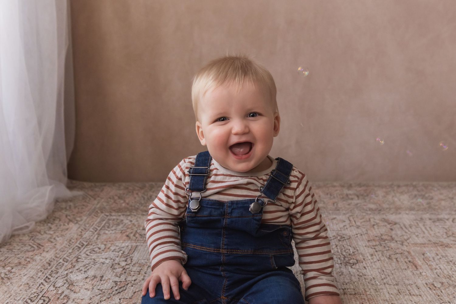 A smiling blonde baby in denim overalls sits on a vintage rug, looking cheerful and playful.