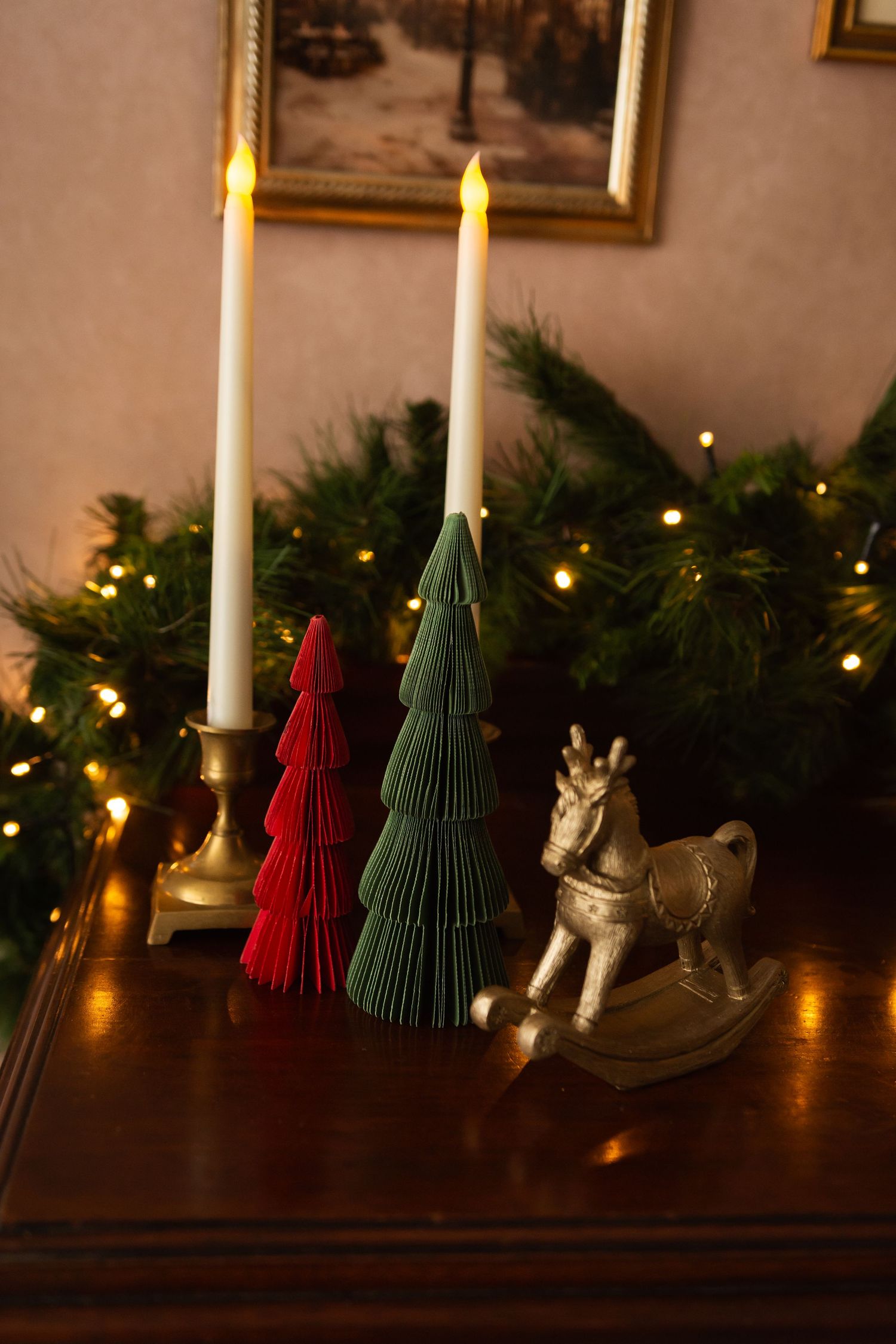 Christmas decor with red and green paper trees, gold rocking horse, lit candles, and festive garland with warm lights.