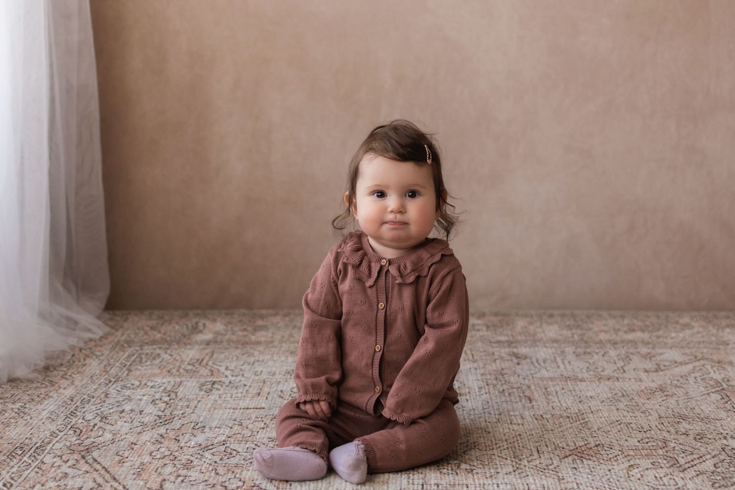 An adorable baby girl in a mauve romper sits independently on a rug in a studio portrait.