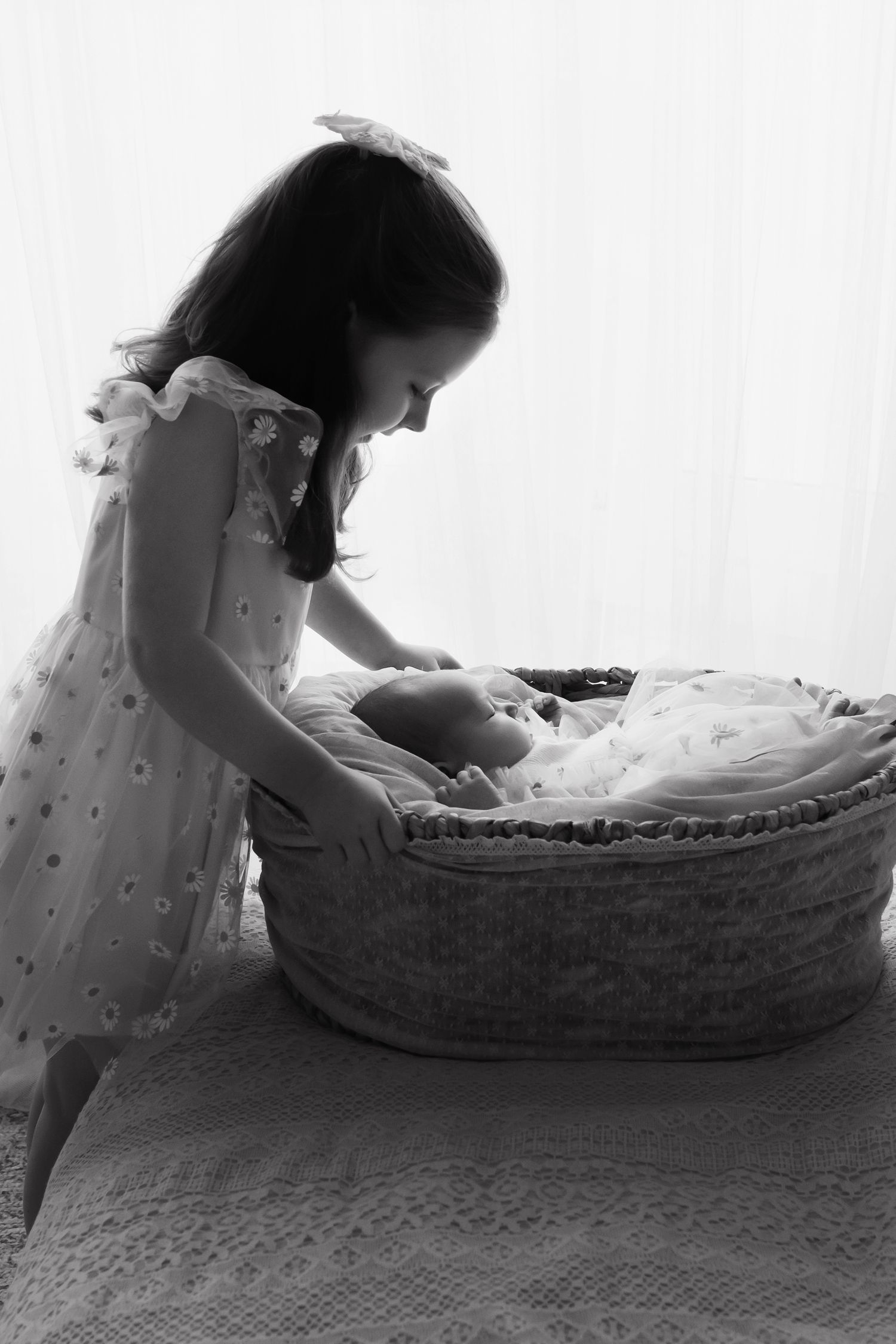 Black and white photo of a little girl in a dress leaning over a wicker Moses basket.