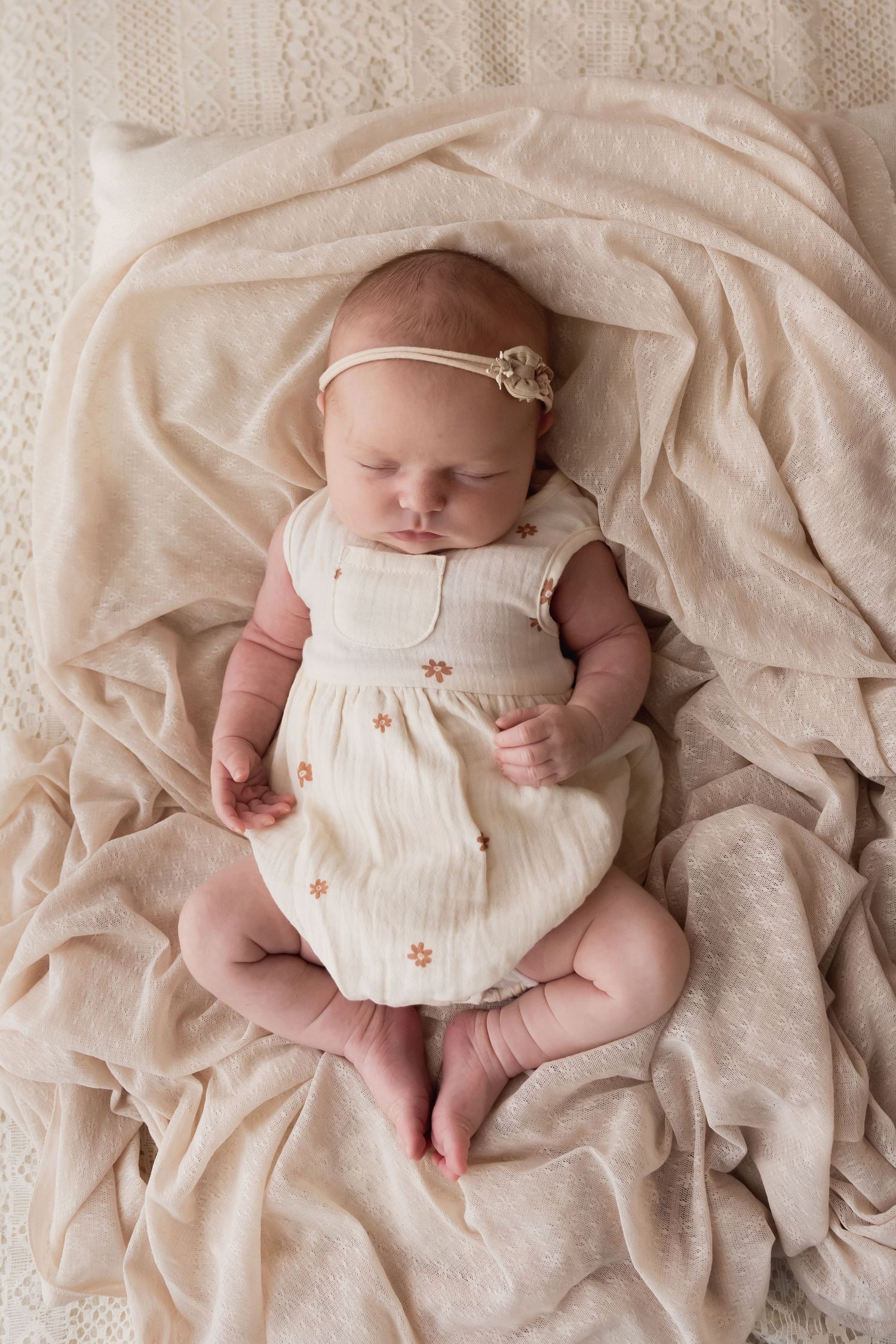 A peaceful newborn baby girl in a cream floral romper sleeps on a soft beige blanket.