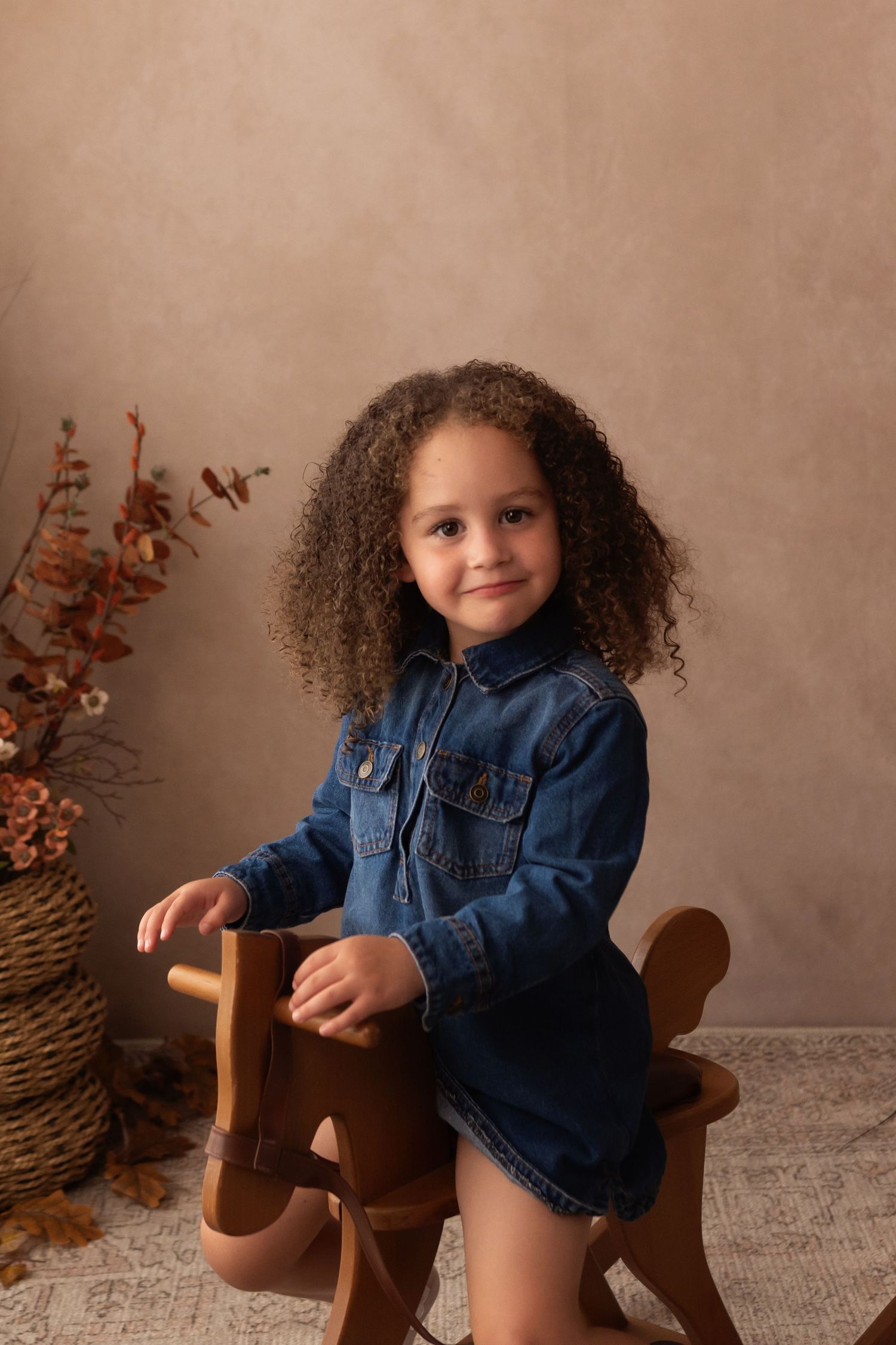 Young girl with curly hair in denim dress sits on wooden rocking horse toy against warm beige studio backdrop.