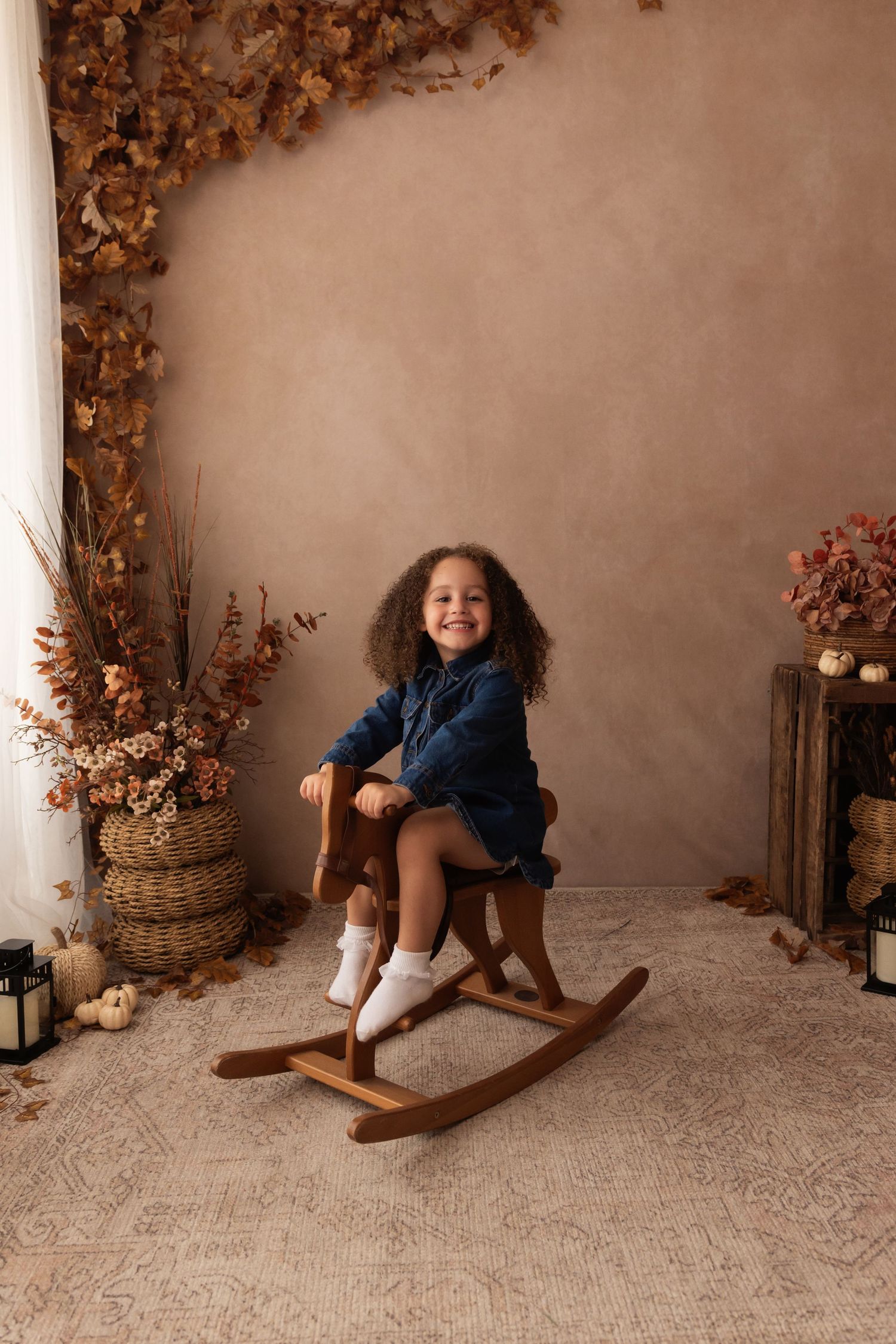 A smiling young girl with curly hair sits on a wooden rocking horse in a fall-themed photo studio setting.