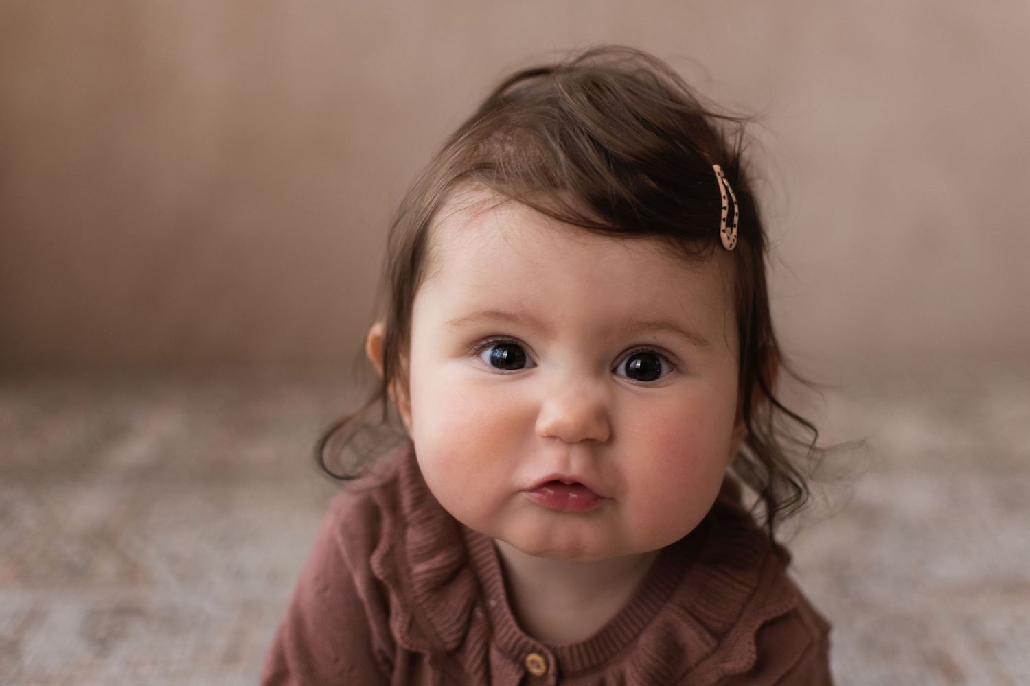 An expressive baby girl in a brown romper stares directly at the camera in a close-up portrait.