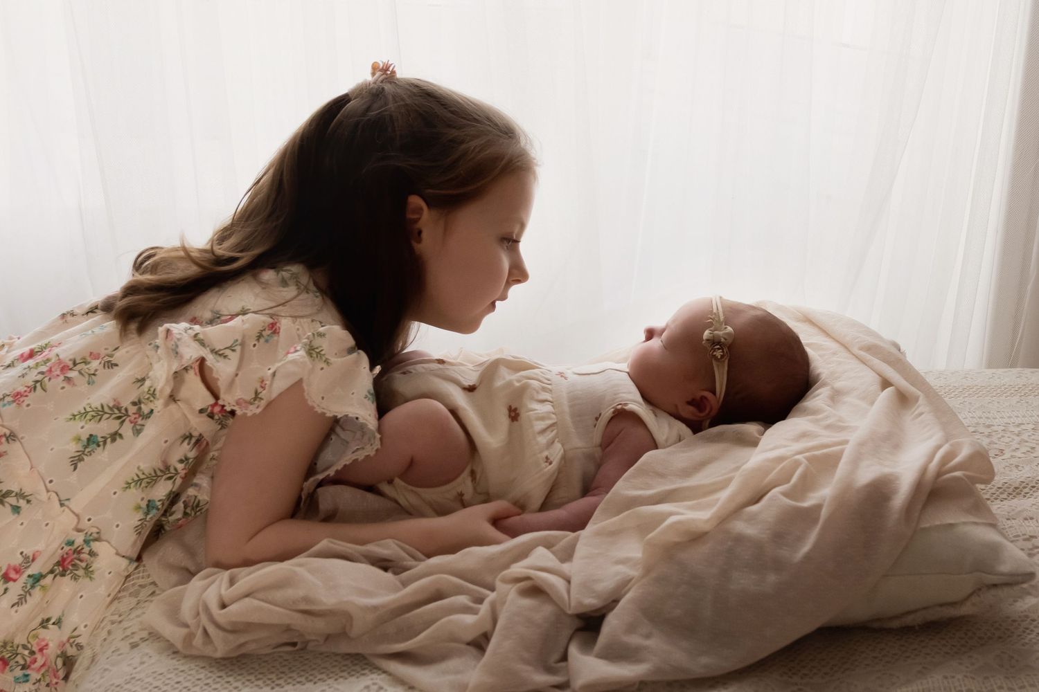 Older sister in floral dress gazing lovingly at sleeping newborn baby.