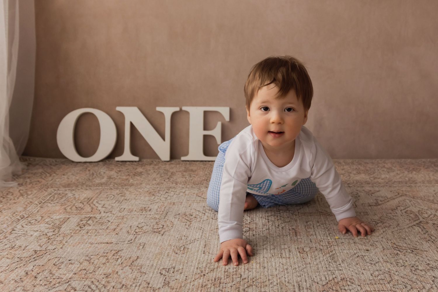 Baby boy in white outfit crawls on a rug toward the camera with large ONE letters behind him.