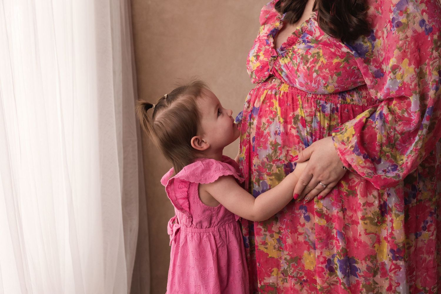 Toddler girl in a pink dress kissing her pregnant mother's baby bump while standing by a curtain.