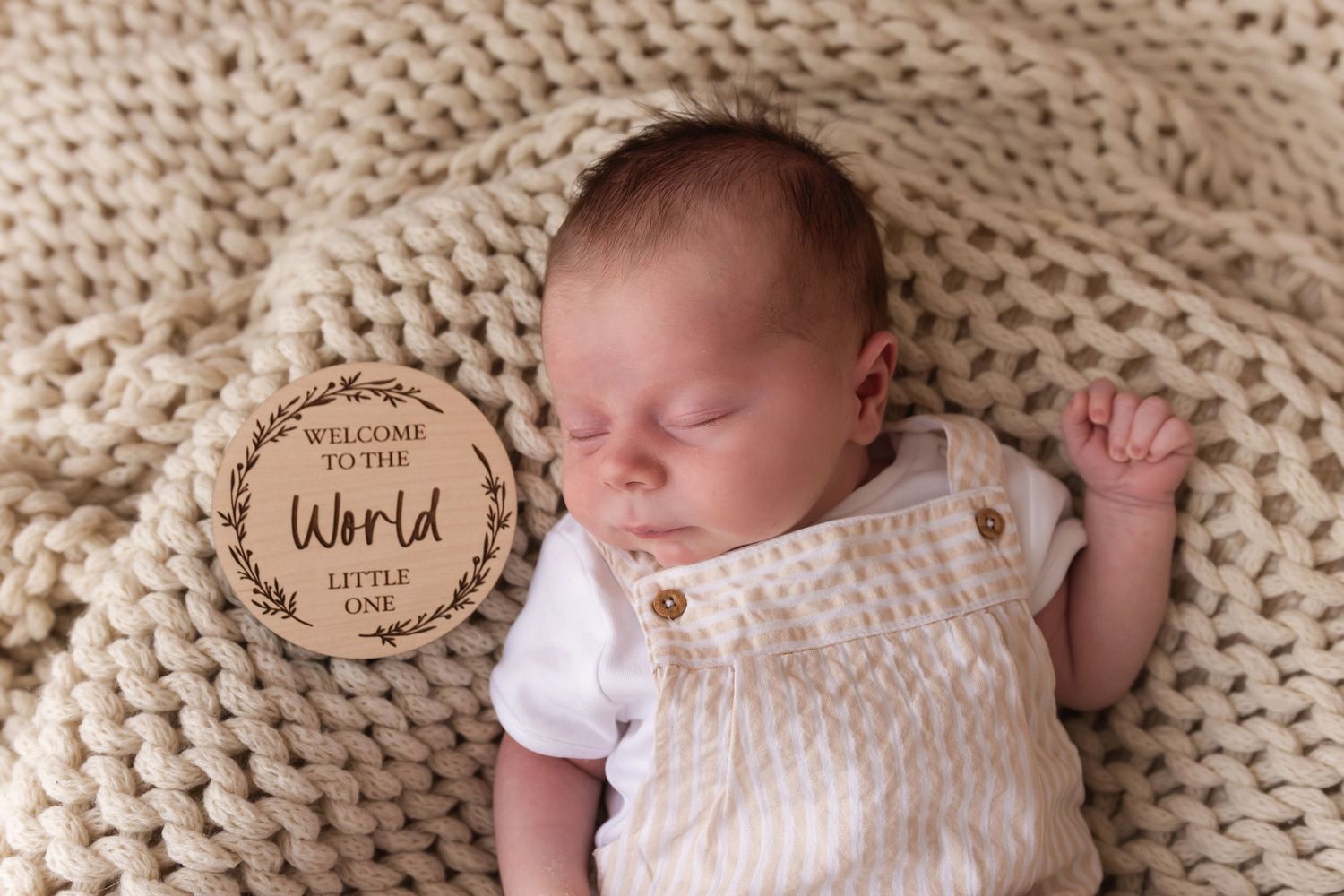 Sleeping newborn baby in cream knit romper beside a Welcome to the World wooden milestone disc.