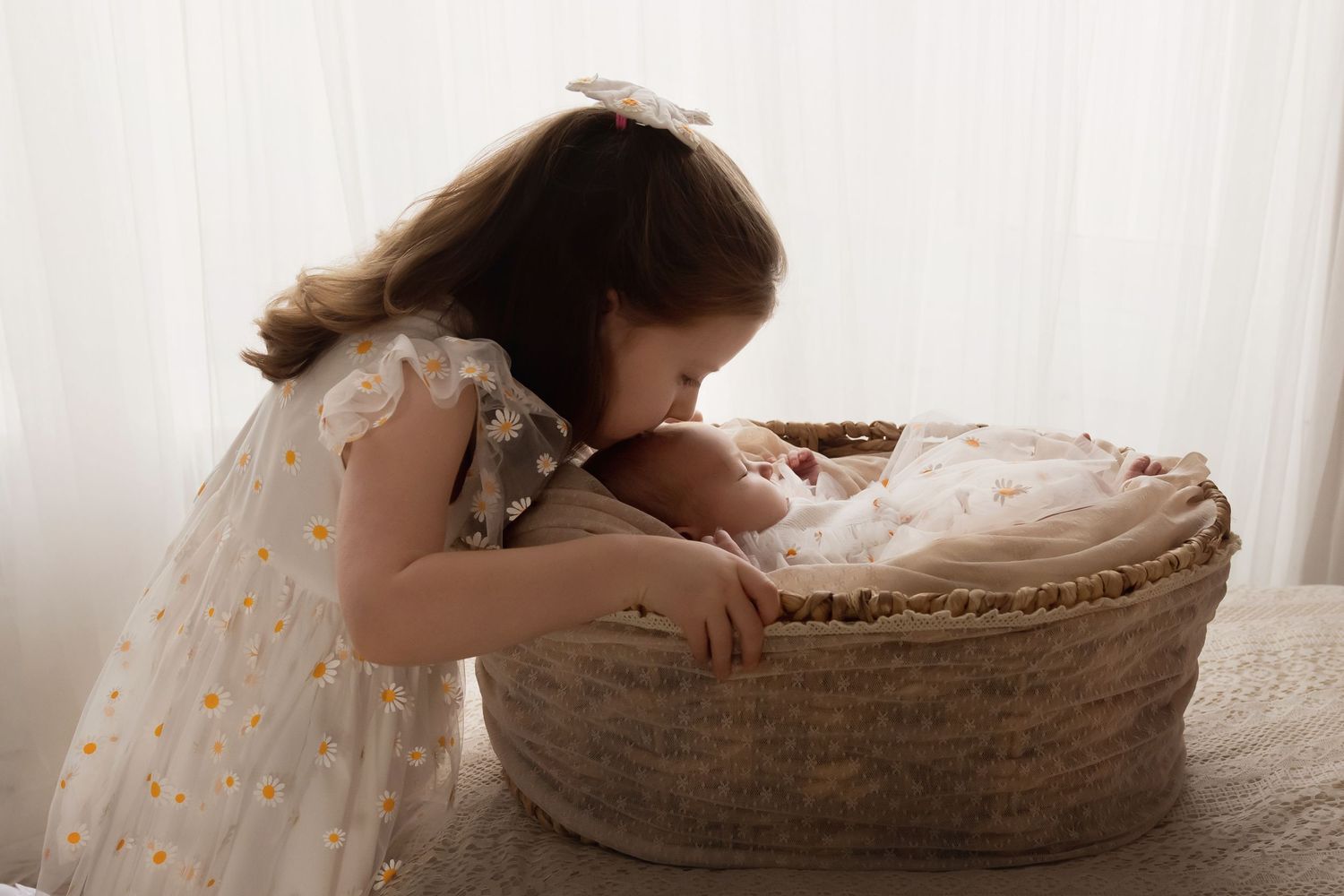 Big sister in daisy tulle dress gently kissing newborn baby in a wicker Moses basket.
