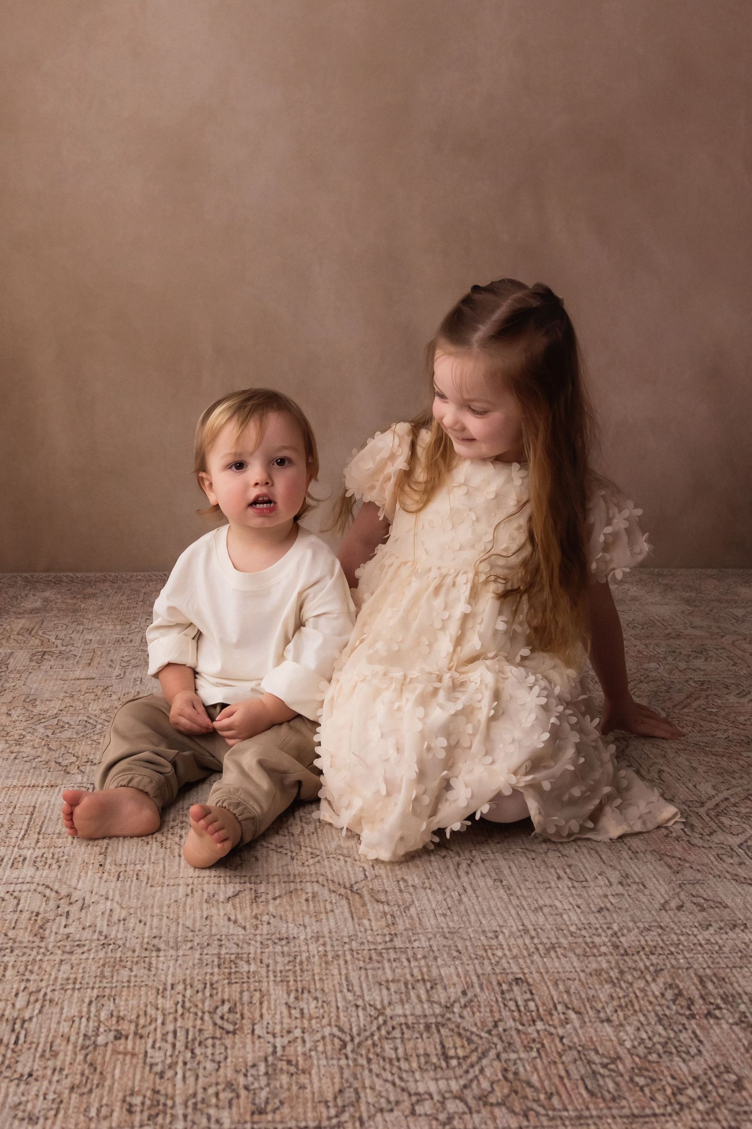 A toddler boy and young girl in elegant cream outfits sit together on a rug against a warm beige backdrop.
