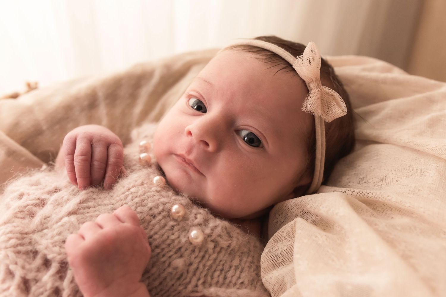 Newborn baby girl in pink knit outfit with pearl details wearing bow headband.
