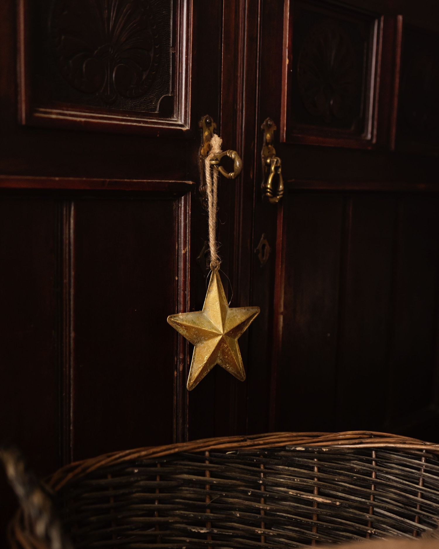 Gold star ornament hanging on rope from dark wooden door panel with wicker basket below.