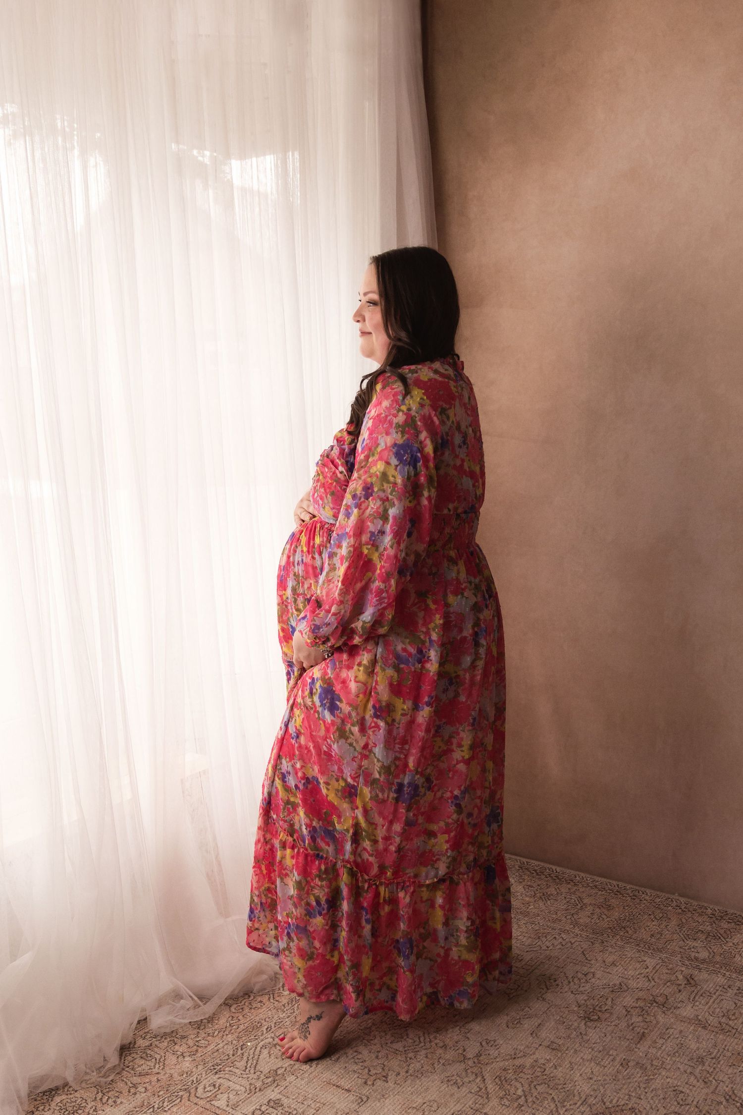 Pregnant woman in vibrant floral maxi dress standing by sheer curtains, cradling her baby bump.