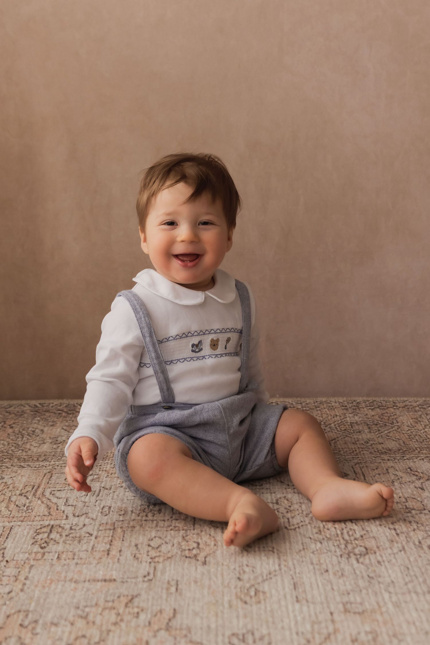Happy smiling baby boy sitting on rug wearing white collared shirt and blue suspender shorts.